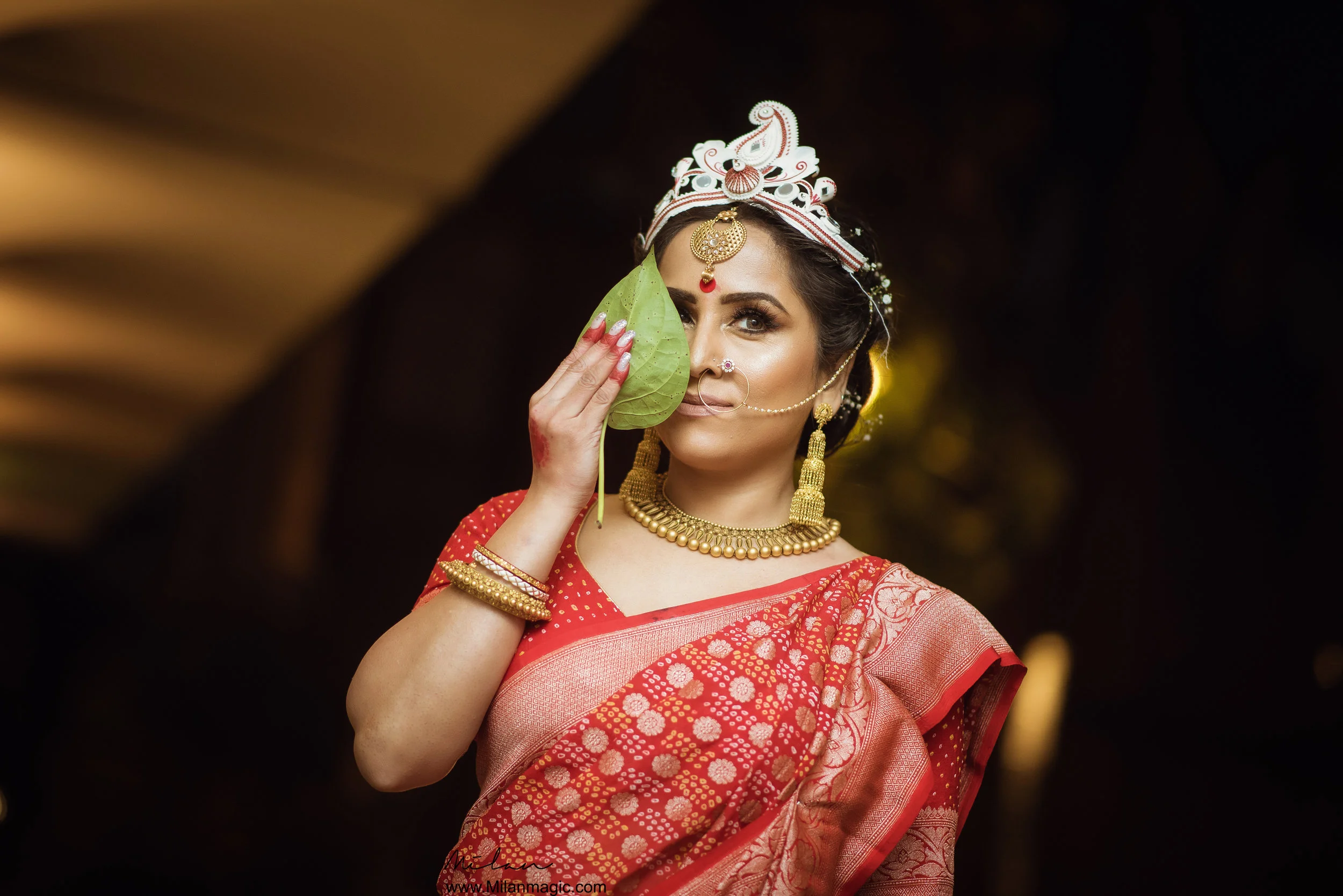 A woman dressed in traditional Indian attire holds a green leaf near her face, with elaborate jewelry and a decorated headpiece, in a dark background.