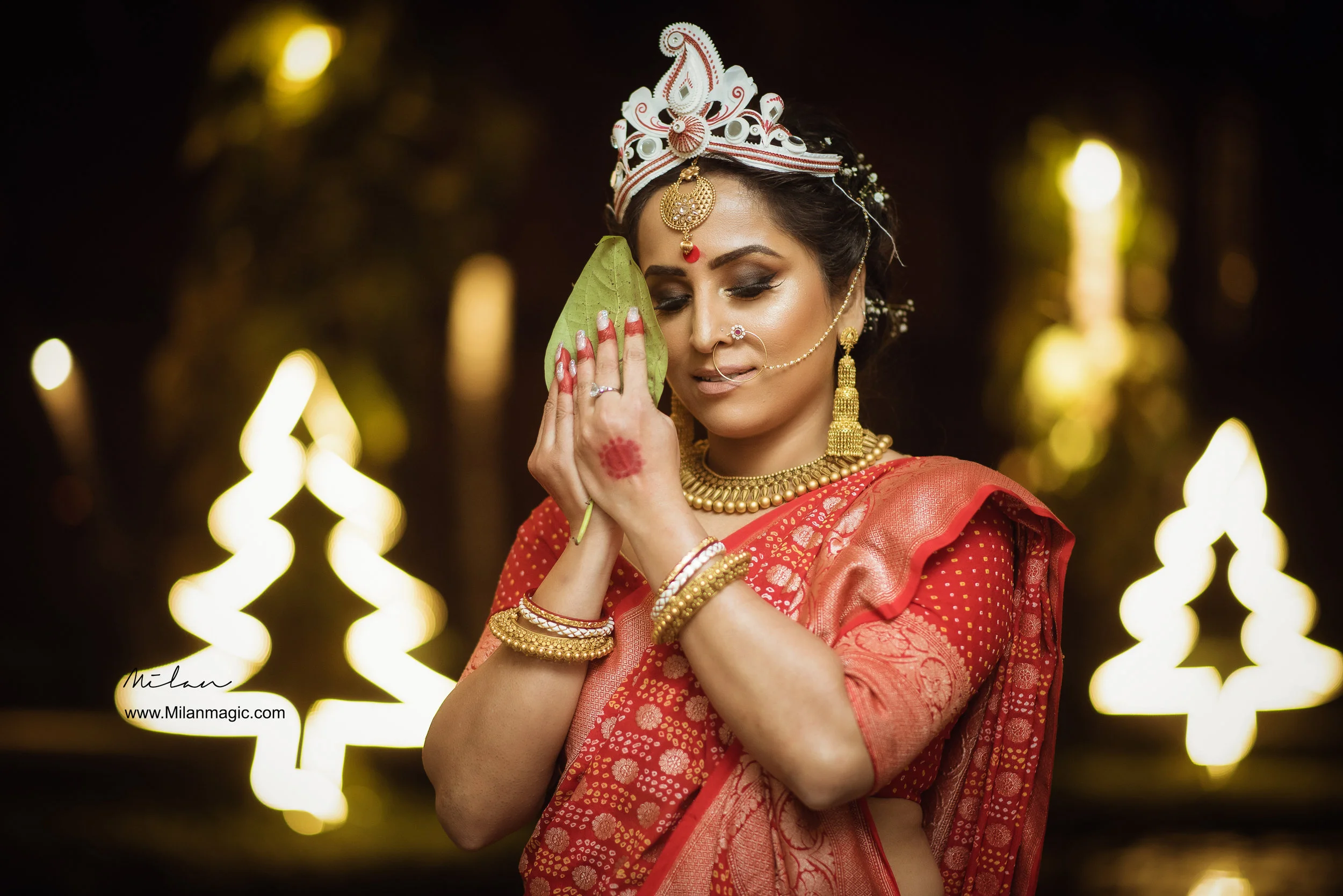 A woman dressed in traditional Indian attire with jewelry, holding a green leaf and embracing it, standing in a festive decorated setting with glowing holiday lights.