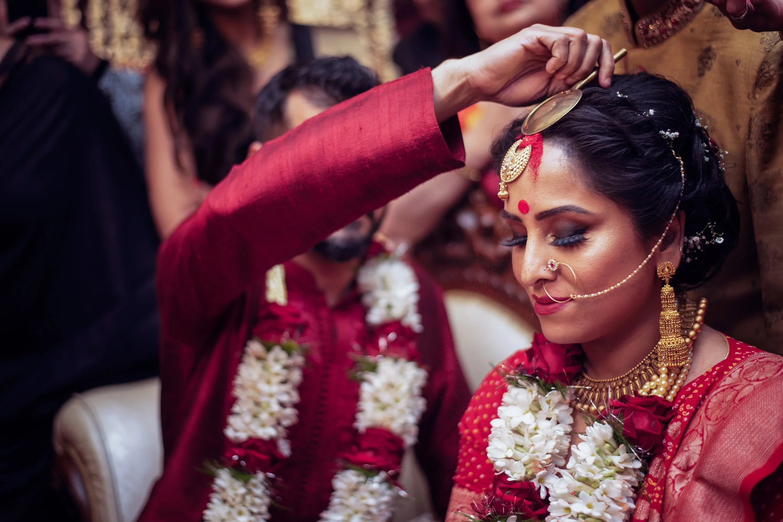 An Indian bride wearing traditional jewelry and a red saree, with a red bindi on her forehead, being blessed or adorned during a wedding ceremony, surrounded by family and friends.