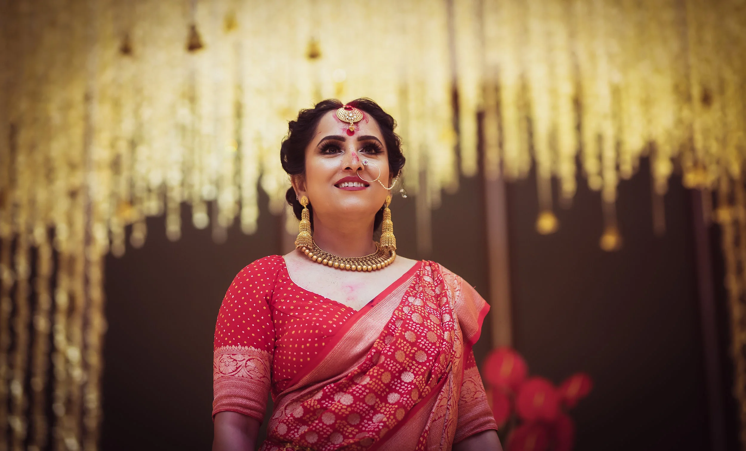 A woman dressed in a traditional red and gold saree with heavy gold jewelry, smiling at a celebration with gold hanging decorations in the background.