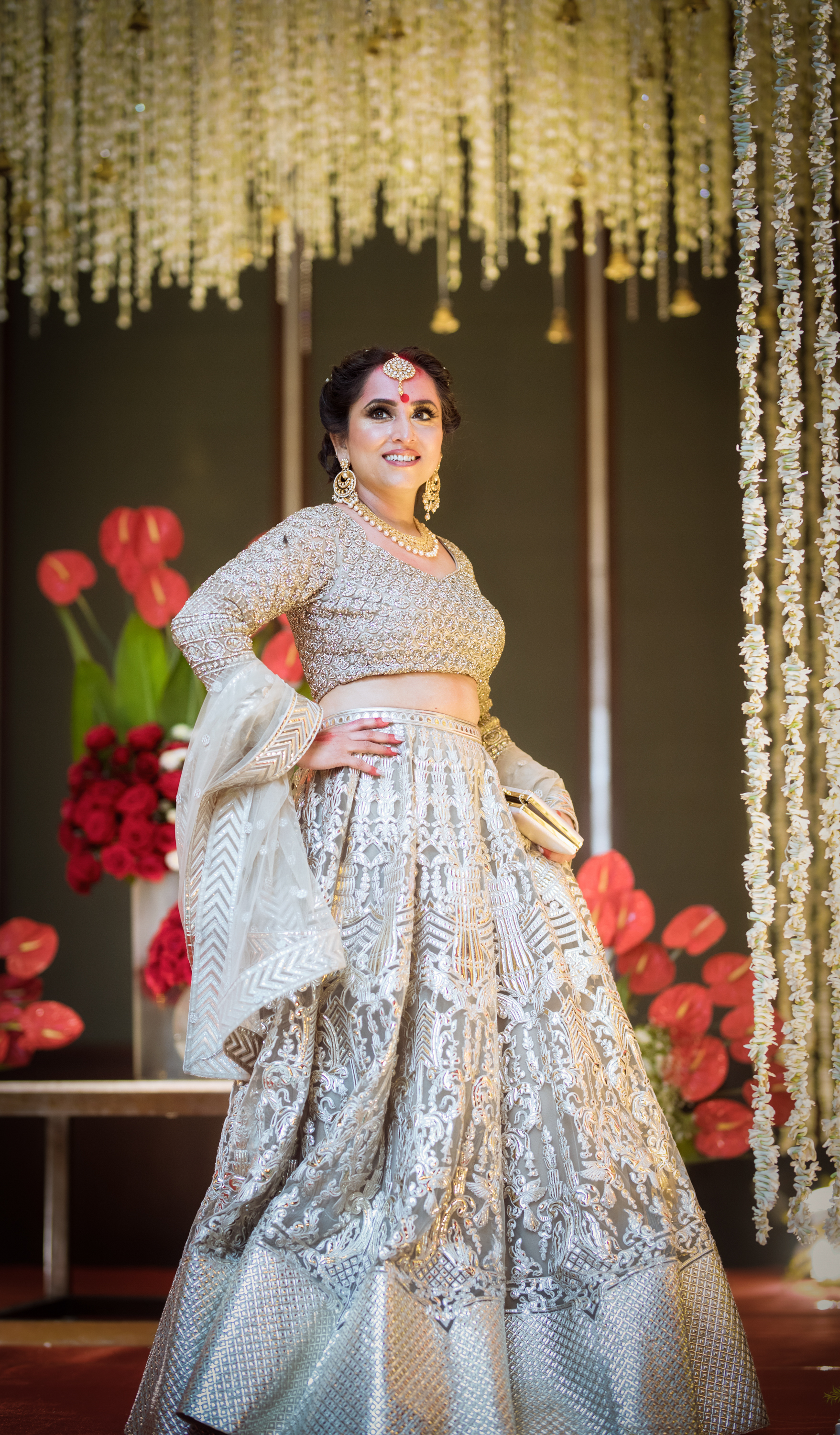 Woman dressed in traditional Indian bridal attire, wearing a silver lehenga with intricate embroidery, gold jewelry, and a headpiece, standing in front of a floral and hanging fabric backdrop with red flowers and white floral curtains.