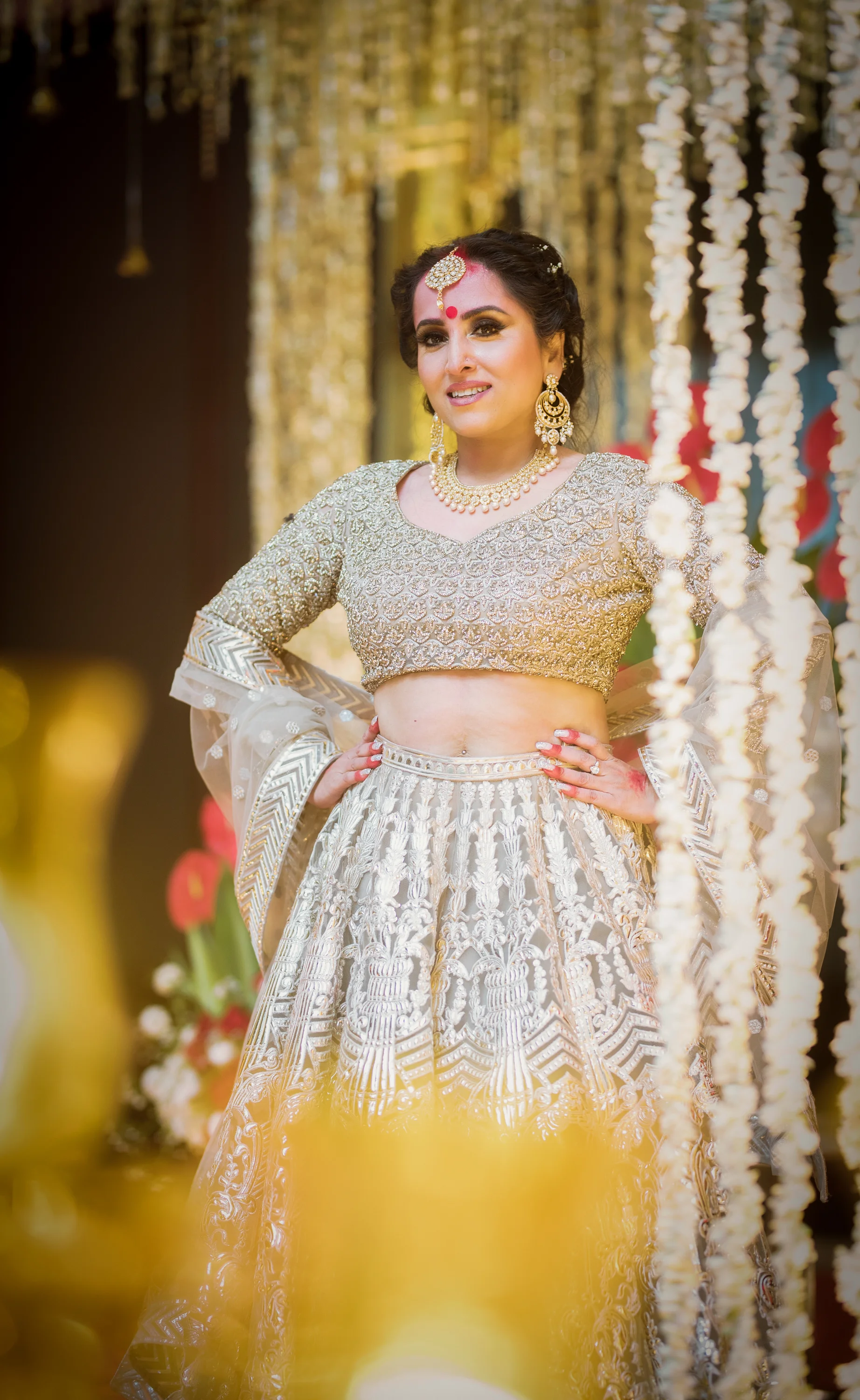 A woman dressed in an ornate traditional Indian outfit, wearing jewelry and standing with her hands on her hips amidst decorative hanging flowers at a celebration or wedding.
