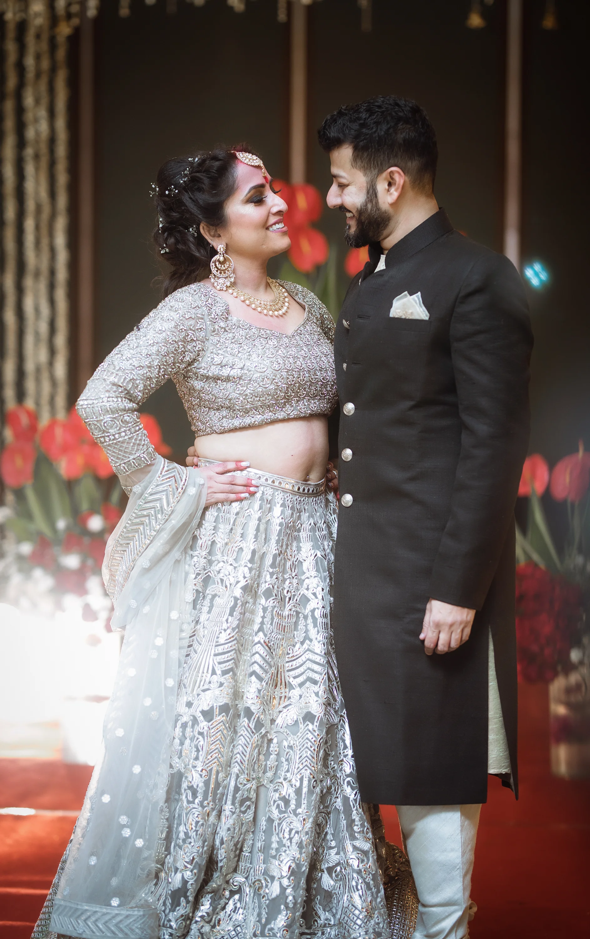 A Bride and Groom smiling at each other during their wedding ceremony, with the bride wearing a traditional silver and white bridal outfit and the groom dressed in a black sherwani.