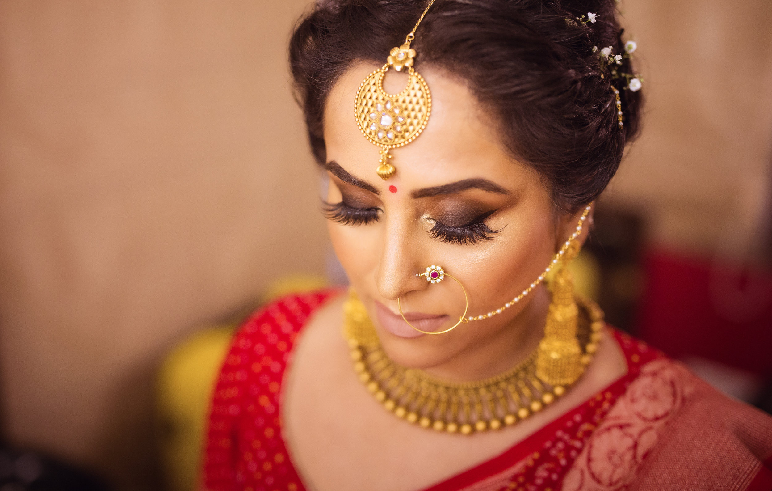 Close-up of a woman dressed in traditional Indian bridal attire with elaborate gold jewelry, including a maang tikka on her forehead, a nose ring connected to her ear, and a choker necklace. She has dark eye makeup and her eyes are closed.