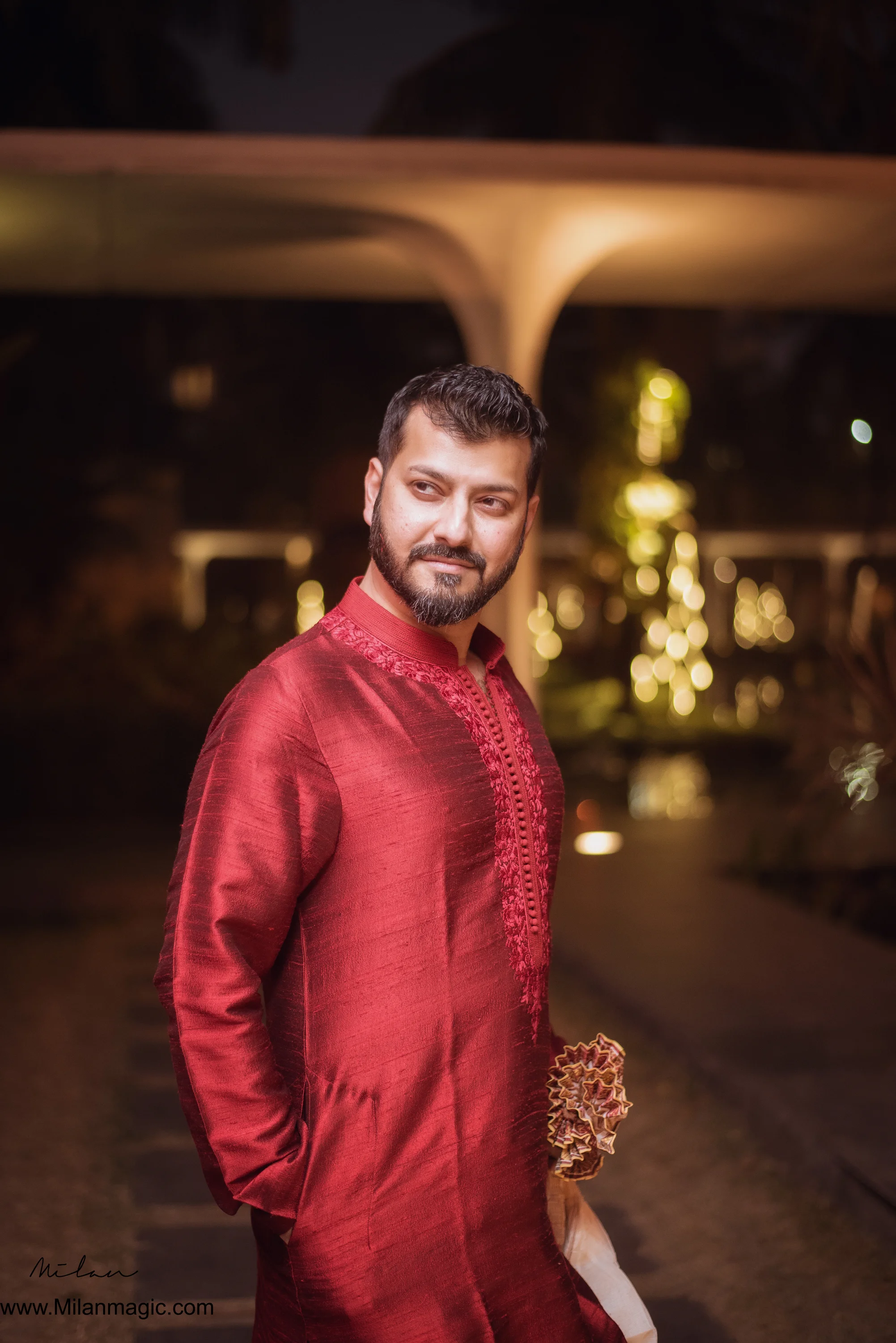 A man in a red traditional outfit standing outdoors at night with blurred lights and trees in the background.