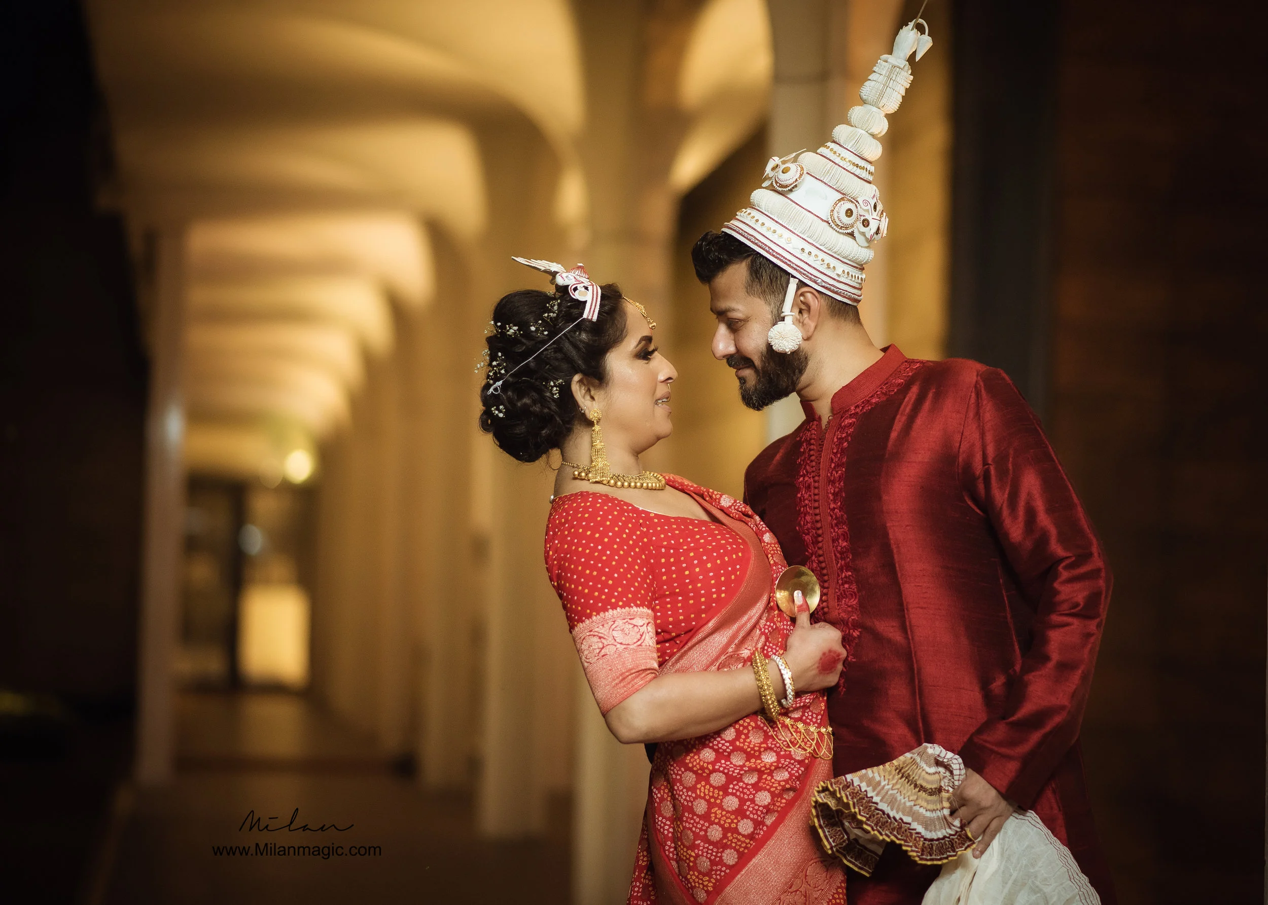 A couple in traditional Indian wedding attire sharing a close moment, the woman in a red saree with gold jewelry and the man in a red kurta with a white traditional headdress, standing in a warmly lit hallway.