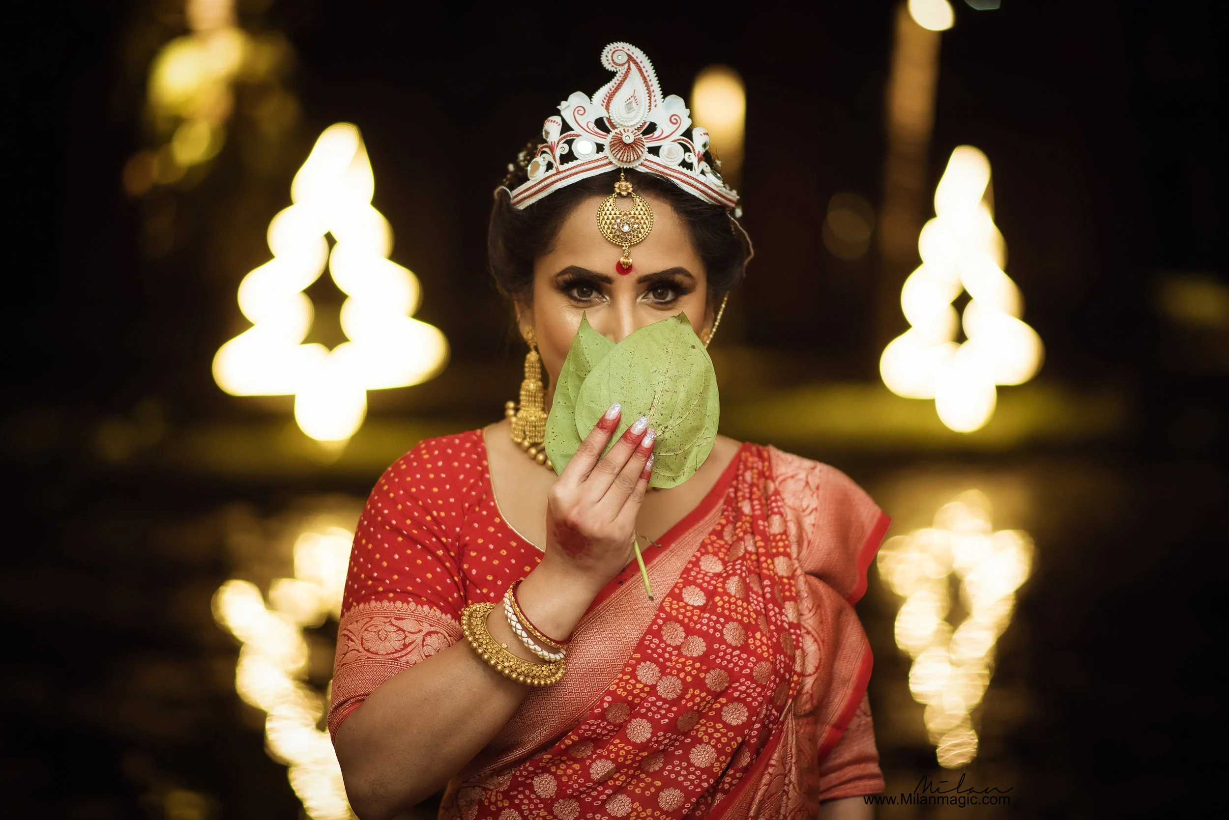 A woman dressed in traditional Indian attire, holding green leaves in front of her face, with a blurred background featuring lights reflecting on water.