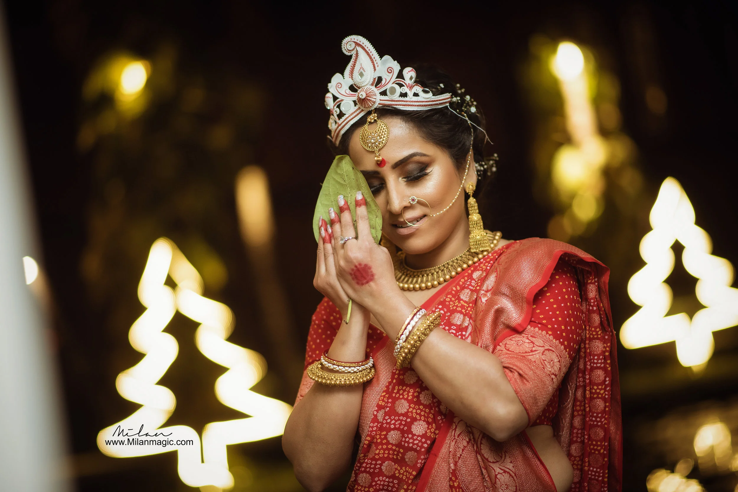 Indian woman in traditional red saree with gold jewelry, holding a green leaf close to her face, with glowing decorations in the background.