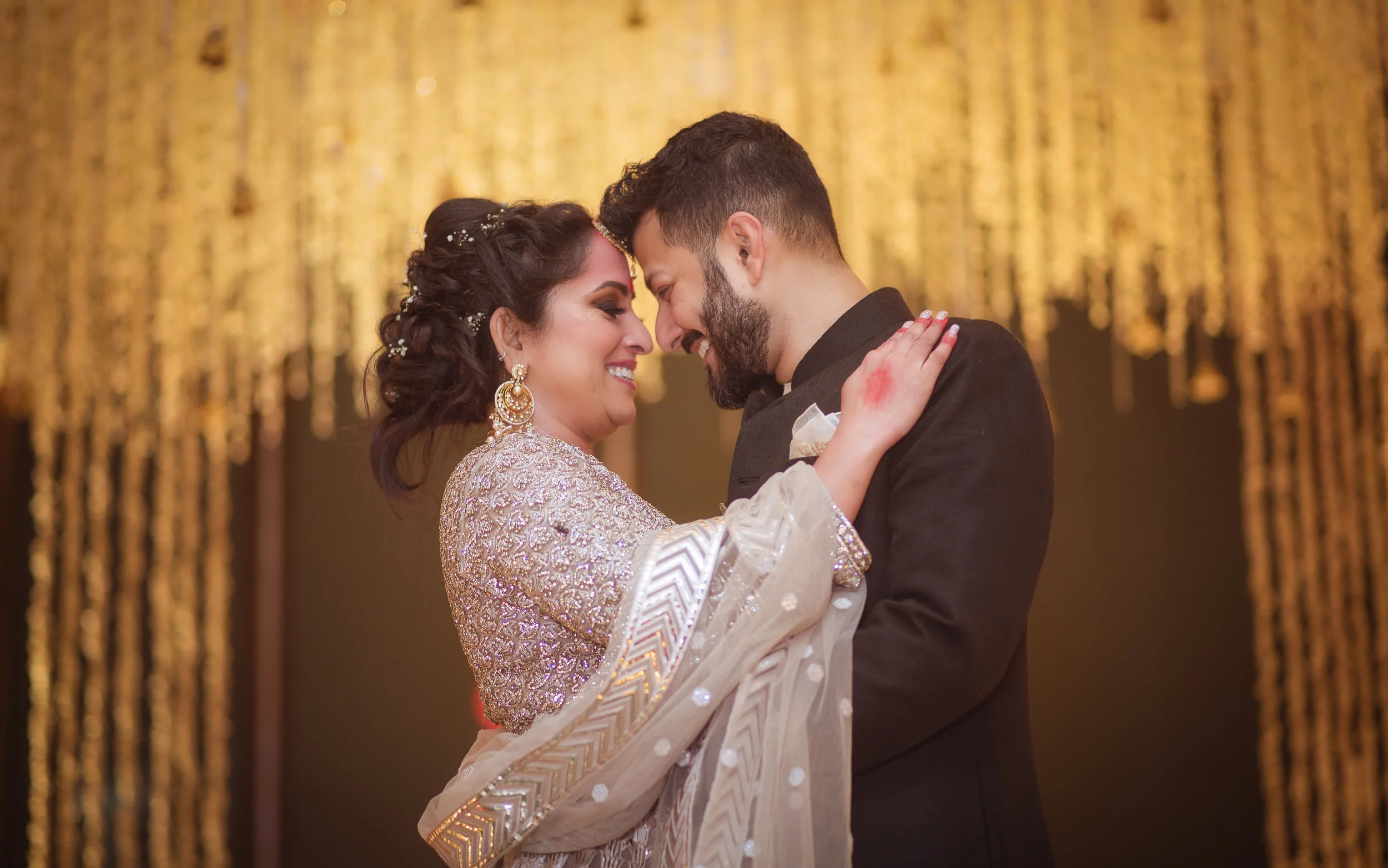 A woman in a traditional embroidered dress and earrings is dancing with a man in a black suit, their foreheads touching and smiling at each other, with golden decorations in the background.