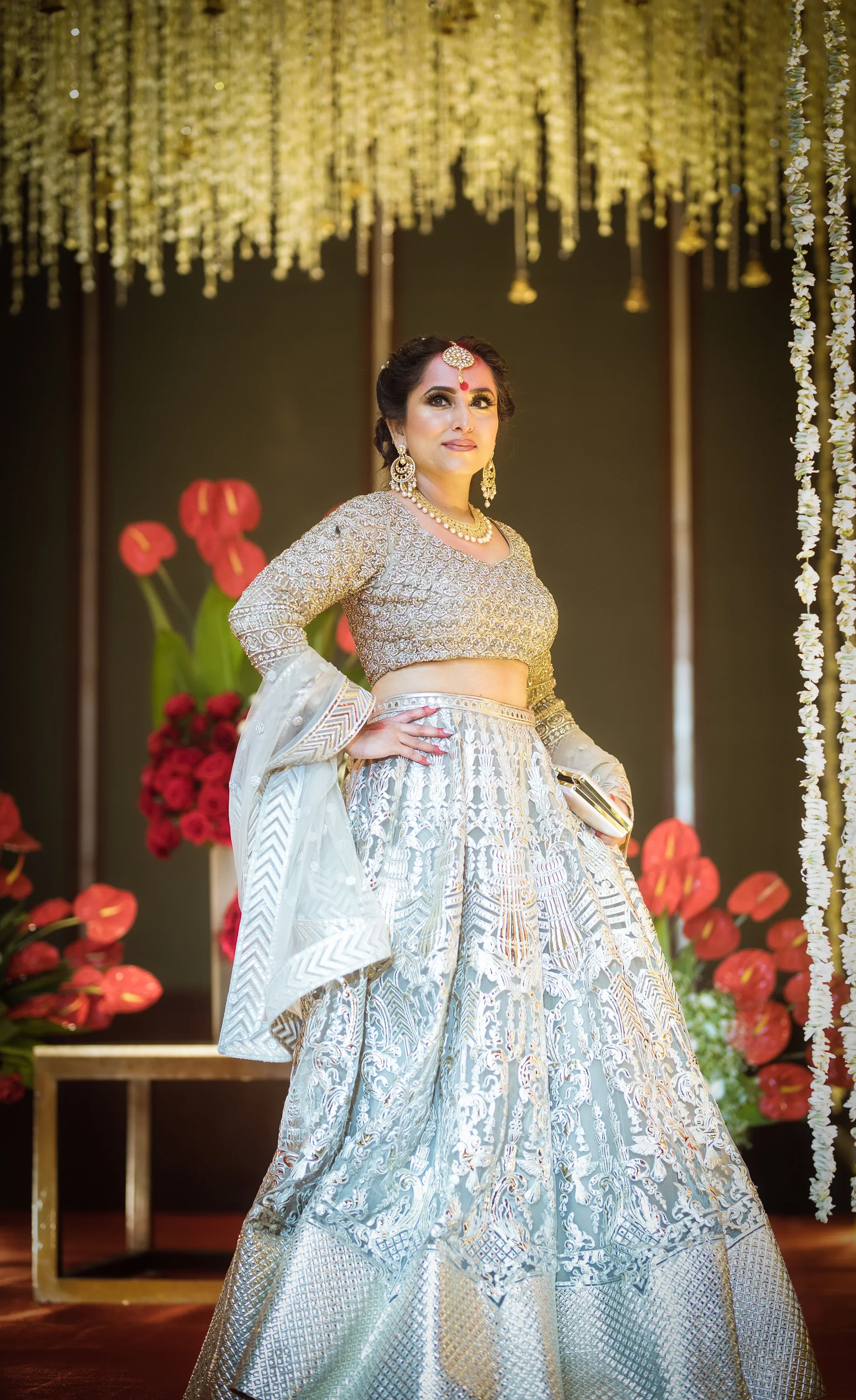 A woman in a traditional Indian outfit, silver and gold embroidered lehenga, standing with her hand on her hip, decorated with jewelry including earrings, necklace, and maang tikka, in a decorated setting with flowers and hanging lights behind her.
