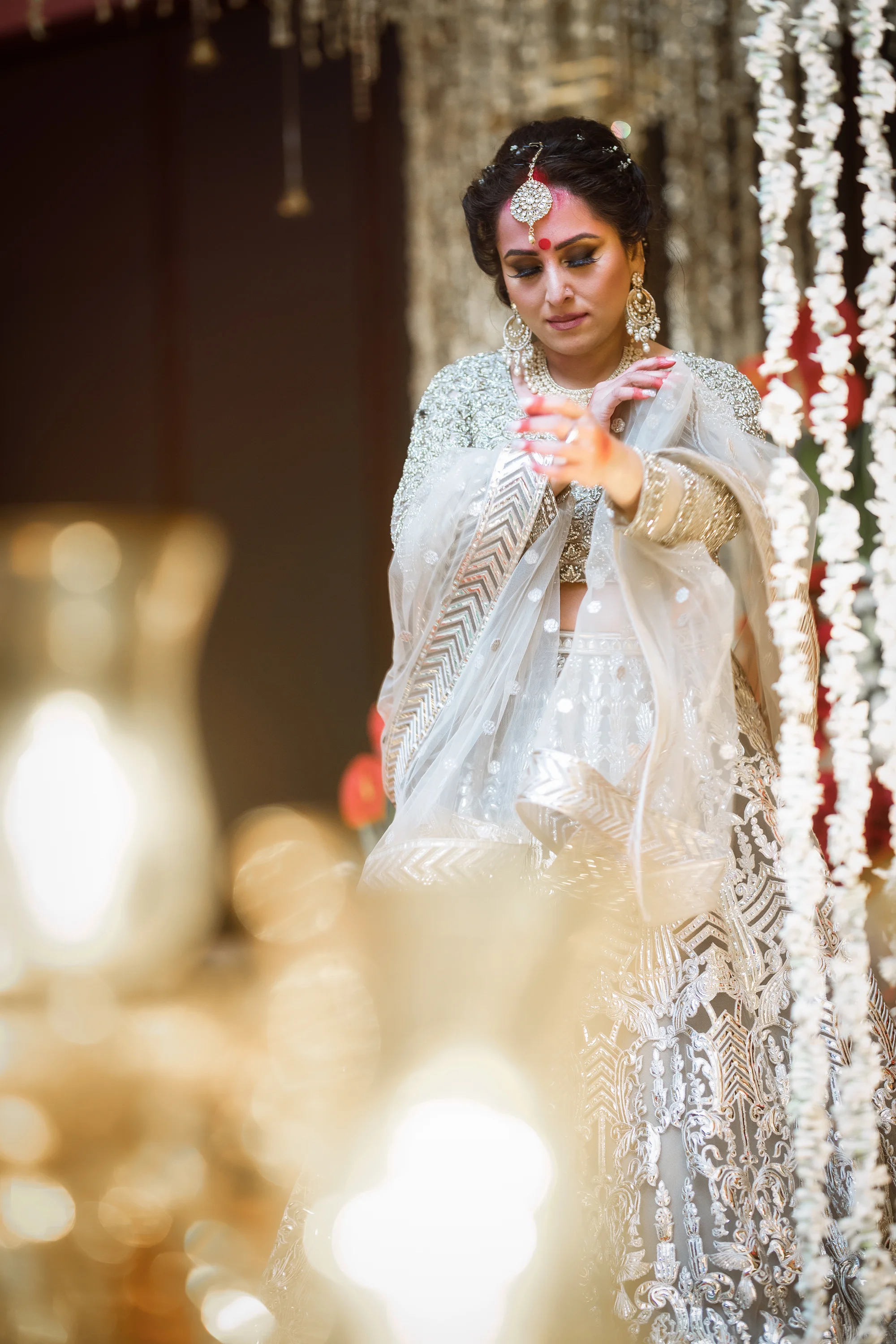 A bride in traditional Indian wedding attire, adjusting her dupatta, surrounded by decorative string flowers and gold accents.