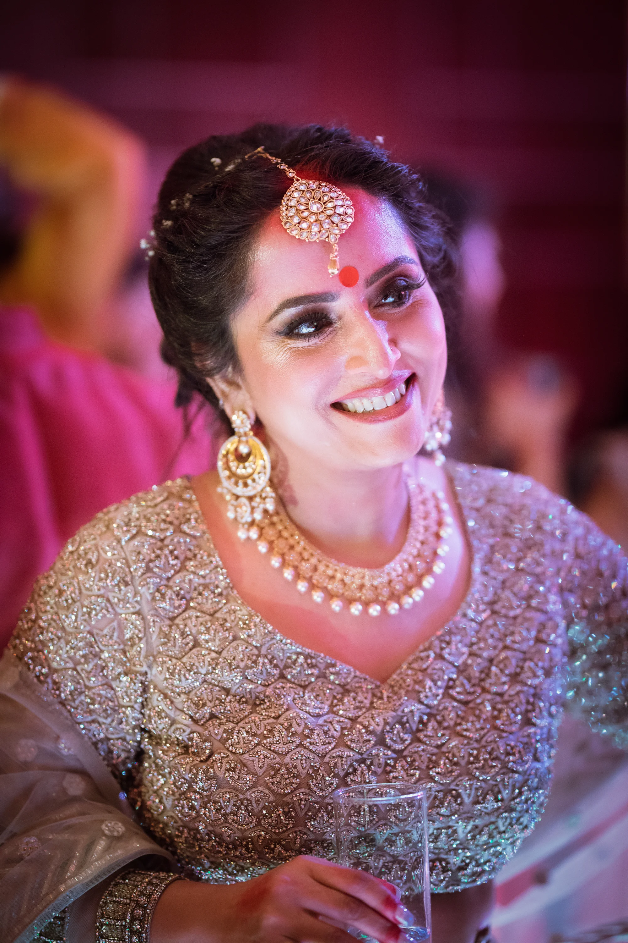 A woman dressed in traditional Indian wedding attire, wearing intricate jewelry, and smiling while holding a glass, at a celebratory event.