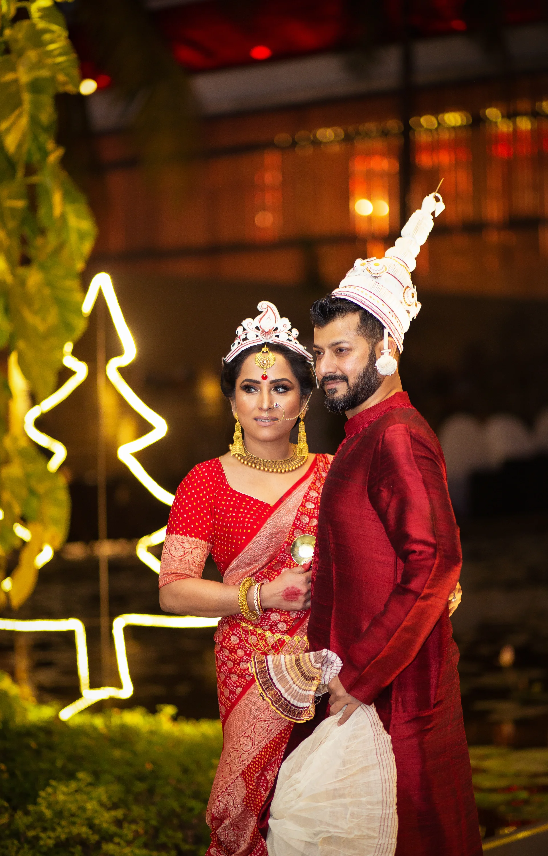 A couple dressed in traditional Indian wedding attire, standing together at night with illuminated decorative lights in the background.