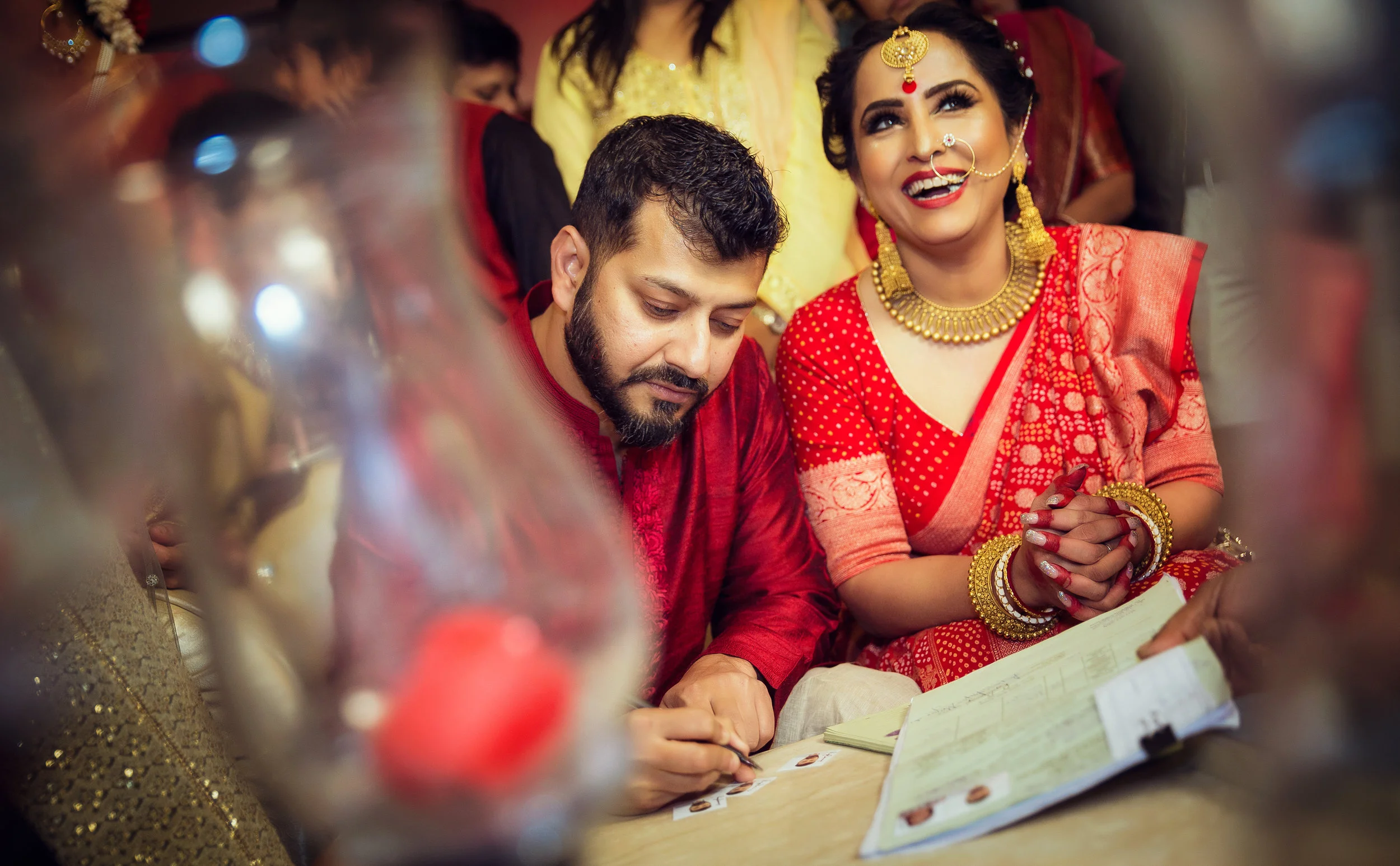 A woman in traditional Indian attire with a red saree and gold jewelry, smiling and looking up, while a man next to her, also in traditional Indian clothing, is signing a document. They are surrounded by others at a celebration.