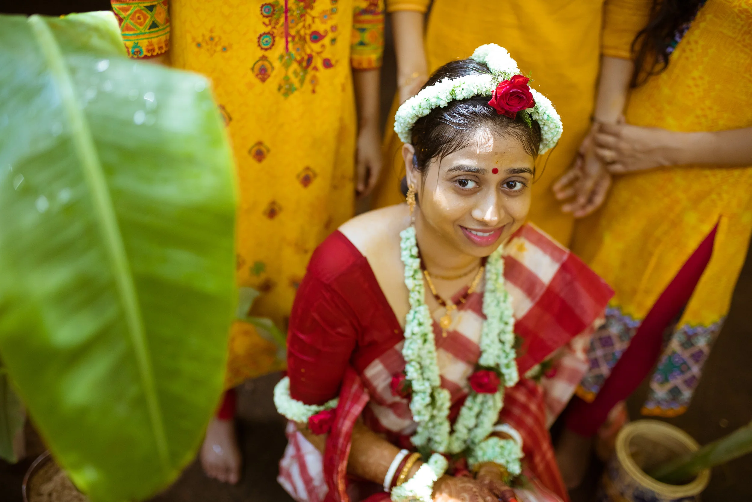 A woman dressed in traditional Indian wedding attire, wearing a red and gold saree, with floral jewelry and a floral headpiece, during a wedding ceremony surrounded by other women in yellow sarees.