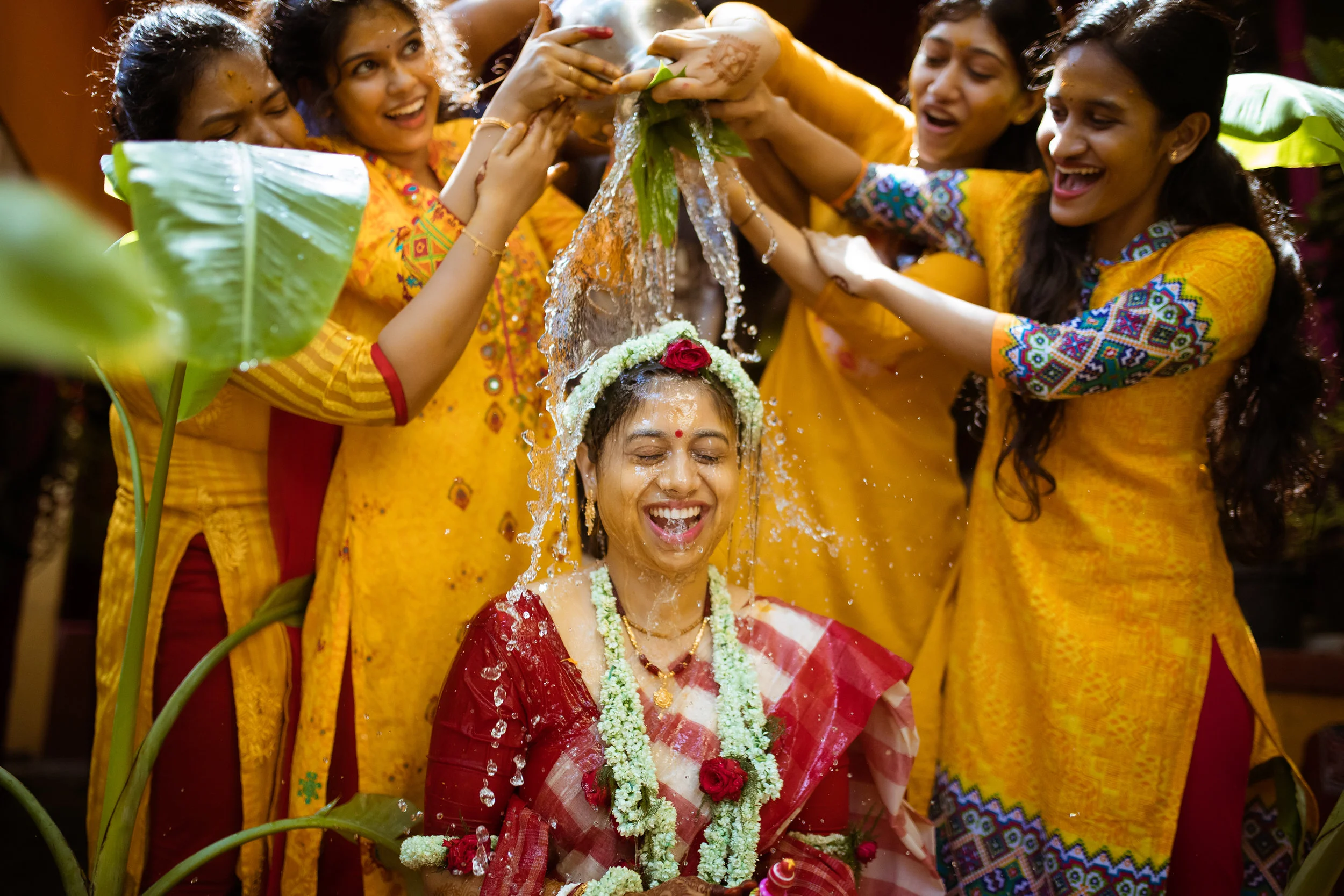 Women in yellow traditional dresses pouring water on a smiling woman in a red and white saree during a festive celebration.