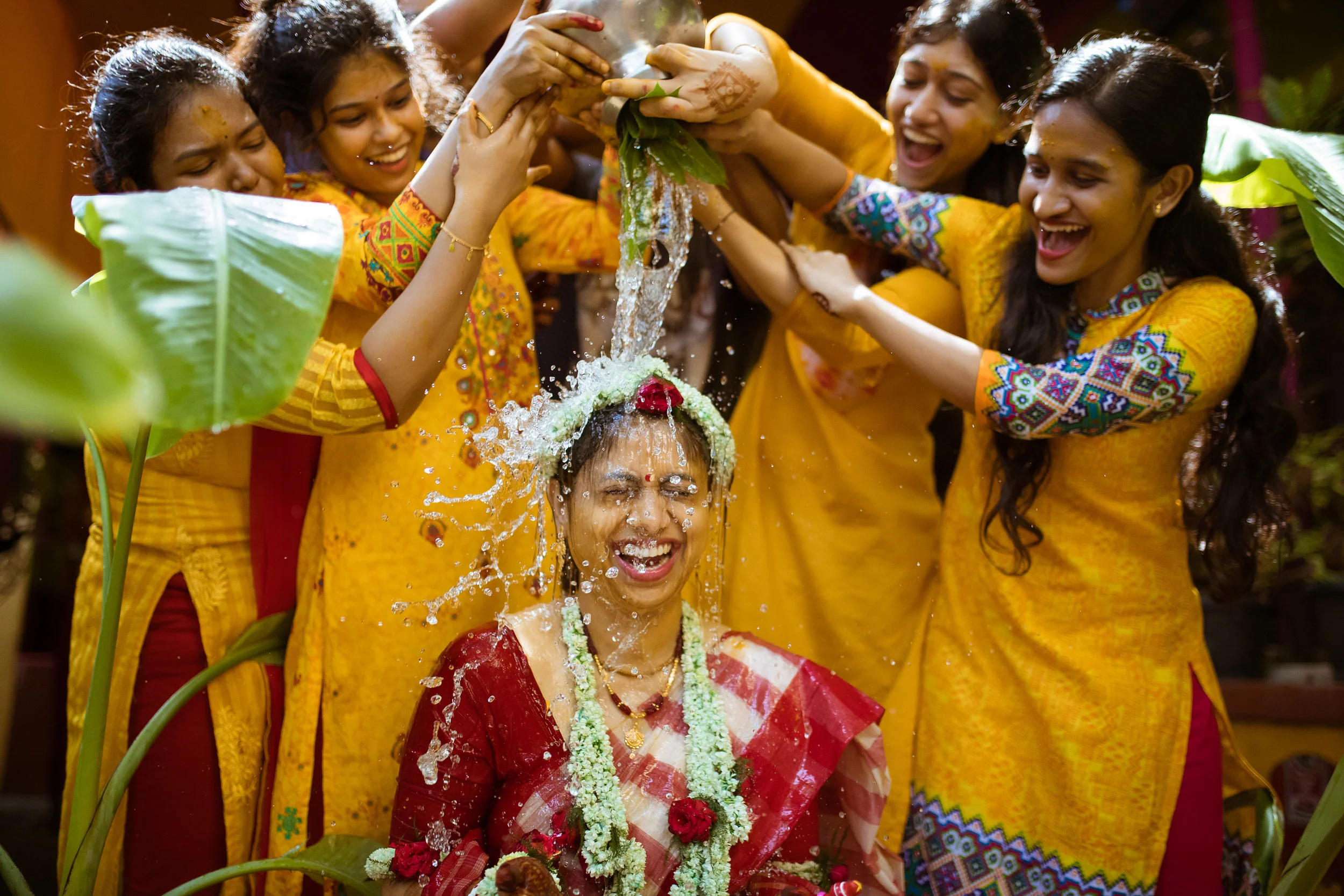 Women celebrating a traditional Indian wedding ritual by showering water on the bride who is wearing red and white saree, floral garlands, and floral headpiece, with other women in yellow sarees around her, in a festive setting.