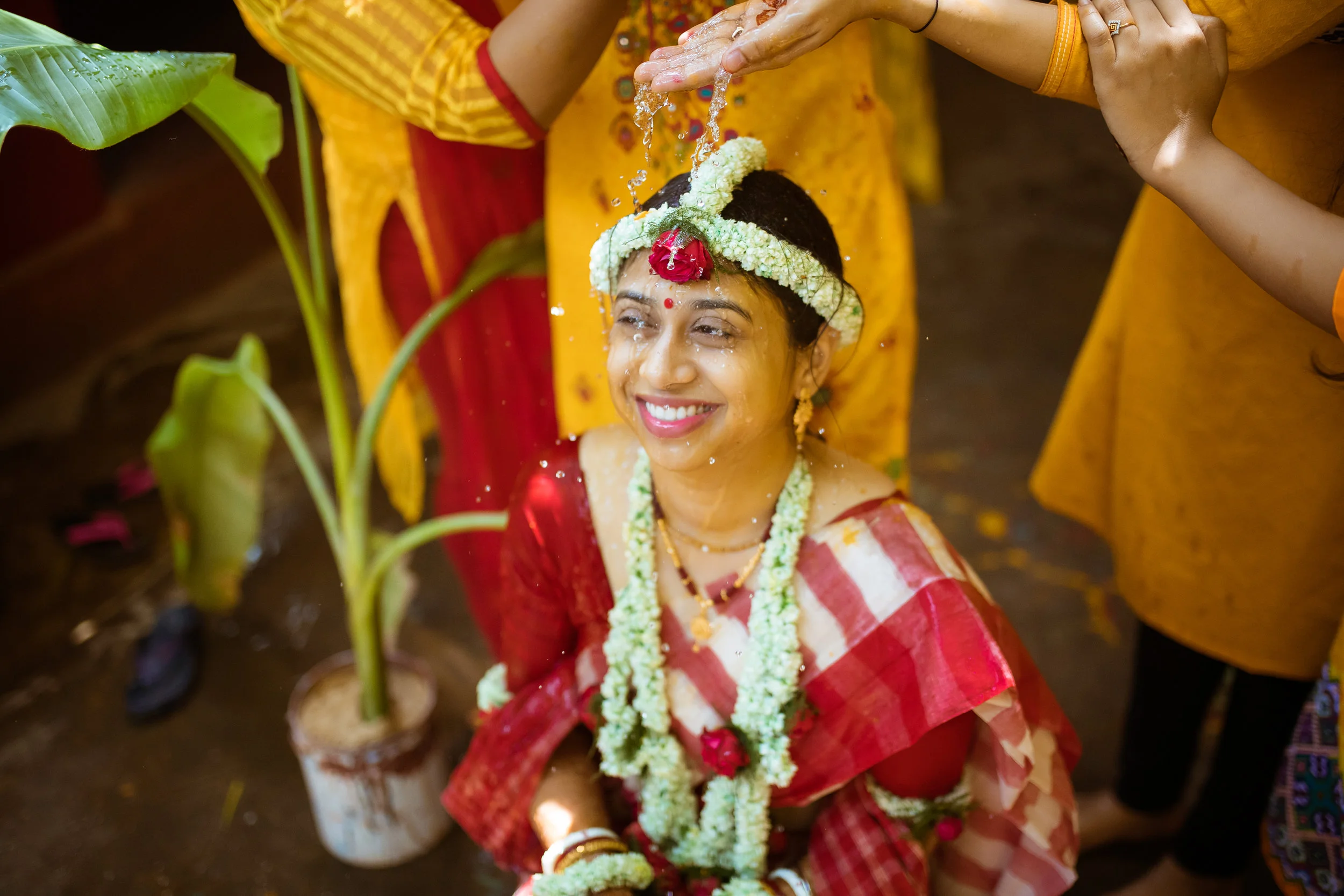 Woman in traditional Indian attire participating in a wedding ritual, water being poured over her head, surrounded by green plants.