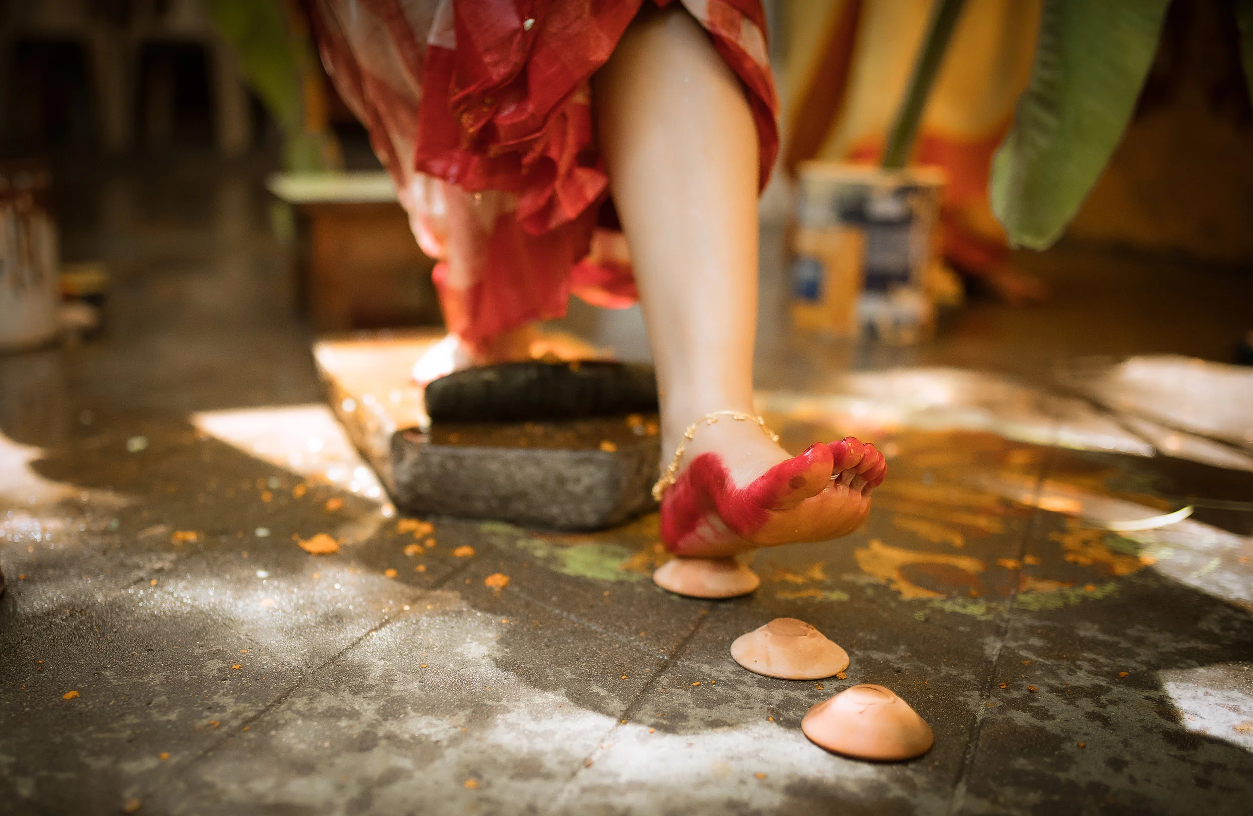 Close-up of a ceremonial statue's foot with a red painted toe, standing on a concrete surface with scattered flower petals and shells. Partially wrapped in a red cloth, with a scene that appears to be part of a religious or cultural ritual.