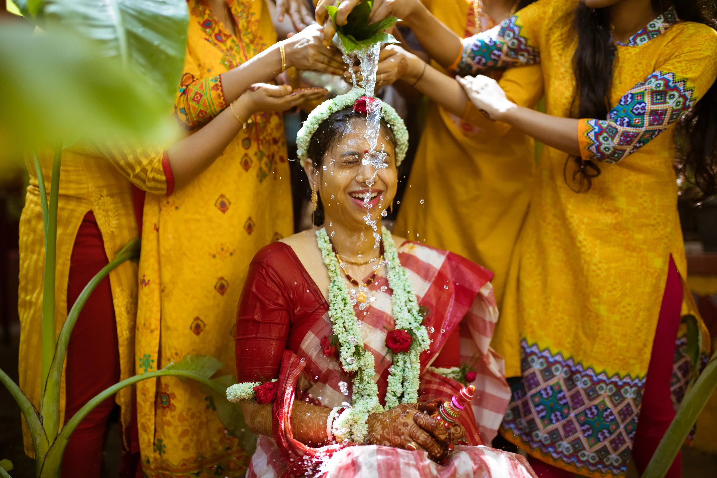 A woman dressed in a red saree with floral jewelry is participating in a traditional Indian wedding ritual, where water is poured over her head by several women dressed in yellow, during a ceremony.