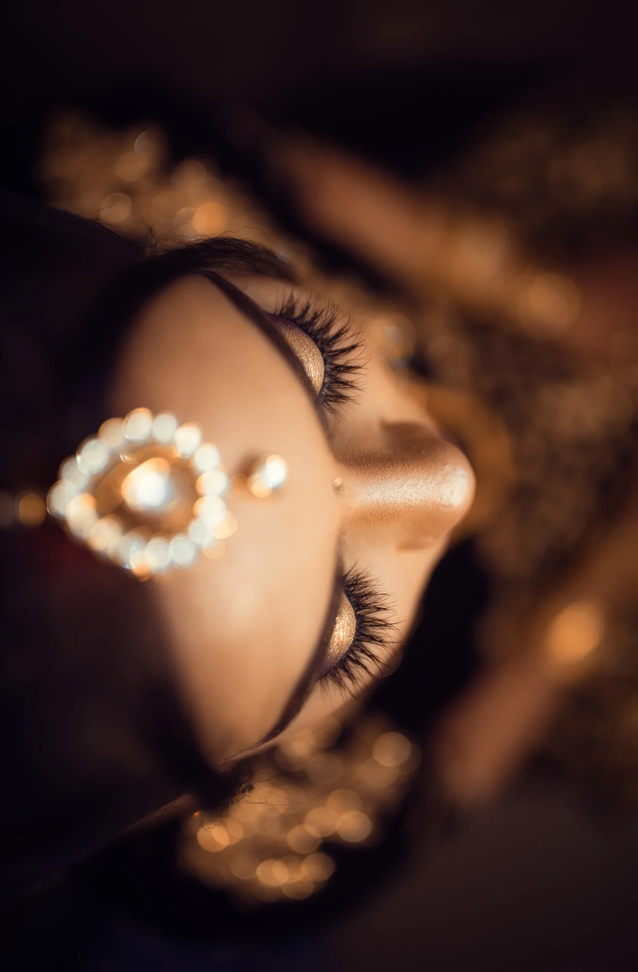 Close-up of a woman's face with gold makeup and jewelry, focusing on her closed eyes with long lashes, gold eyeshadow, and a jeweled nose ring.