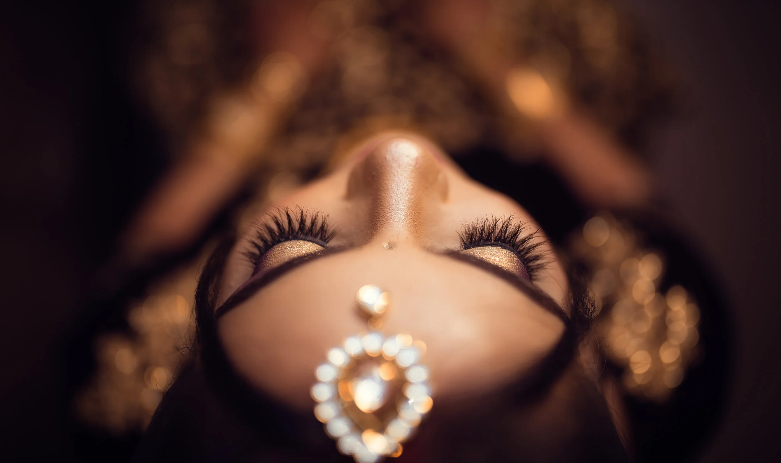 Close-up of a woman's face from a downward angle, showcasing her makeup and jewelry, with her eyes closed and adorned with shimmering eye shadow and long lashes, and a decorative piece on her forehead.