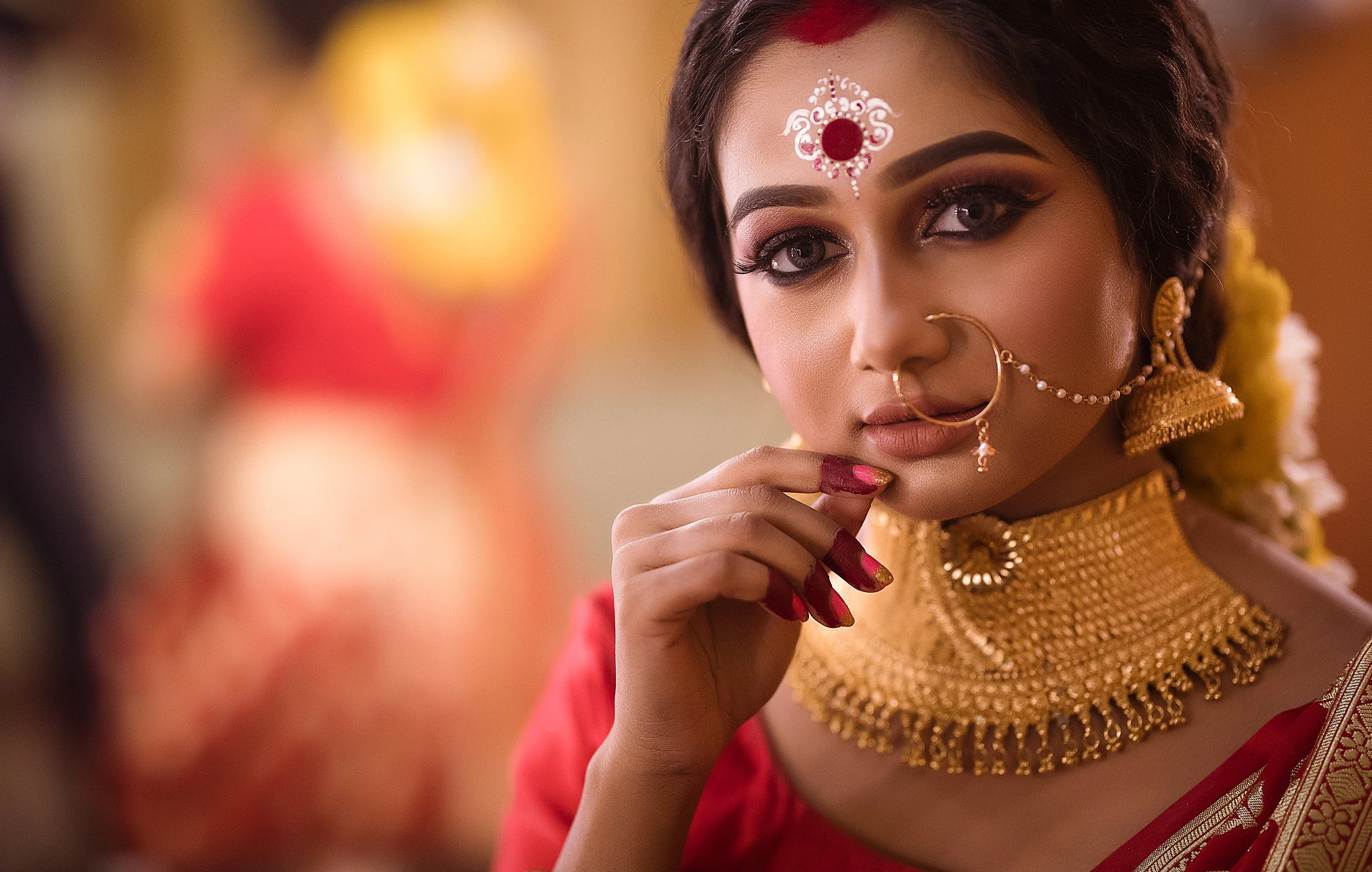 Close-up of a woman dressed in traditional Indian attire with intricate gold jewelry, including a large nose ring connected to her ear, a choker necklace, earrings, and forehead adornment. She has detailed eye makeup and red nail polish, and is looki