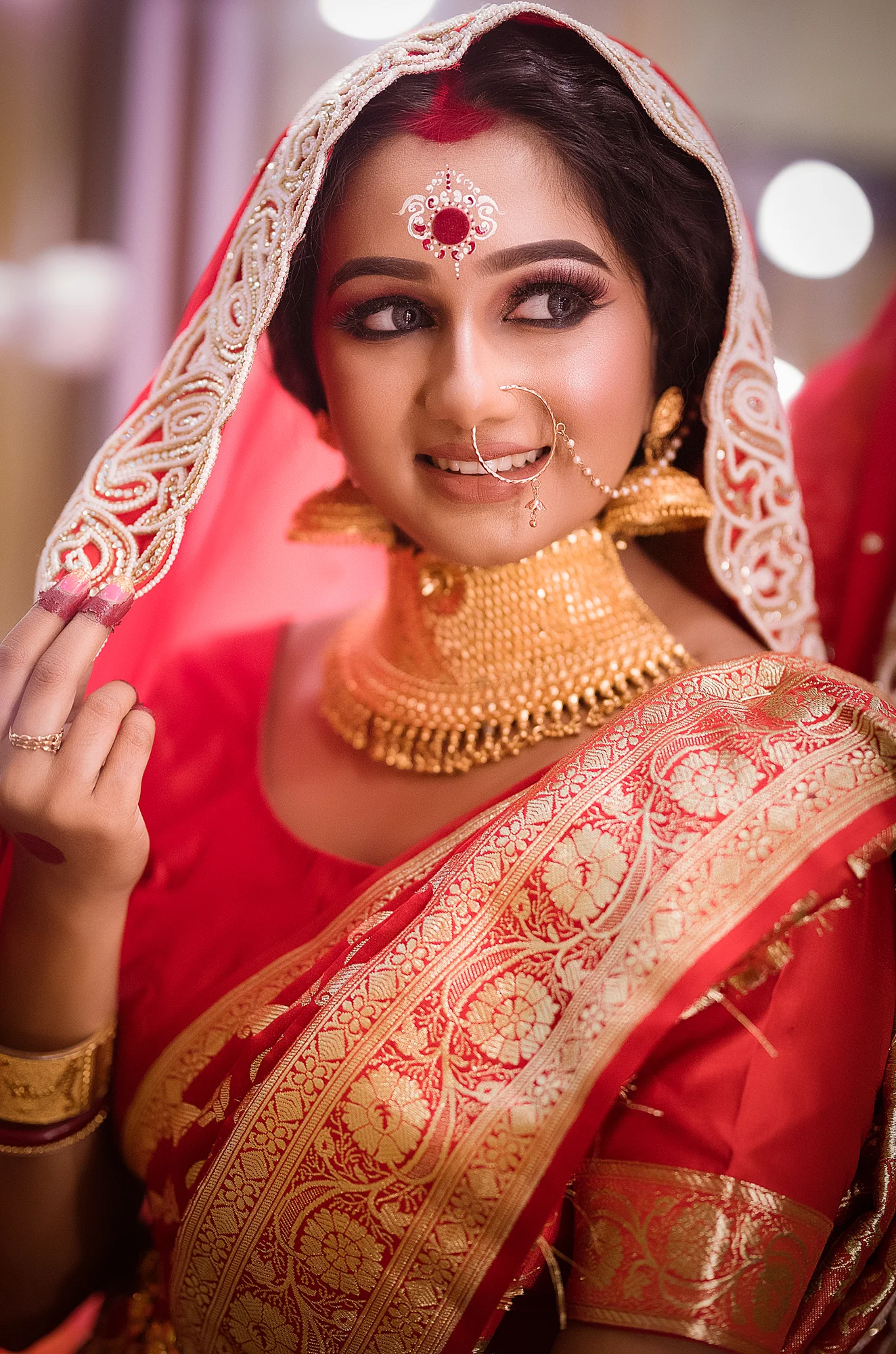 A woman dressed in traditional Indian bridal attire, wearing elaborate gold jewelry including a nose ring, necklace, earrings, and a head covering with intricate embroidery. She has makeup and a decorative red and white bindi on her forehead. She is 