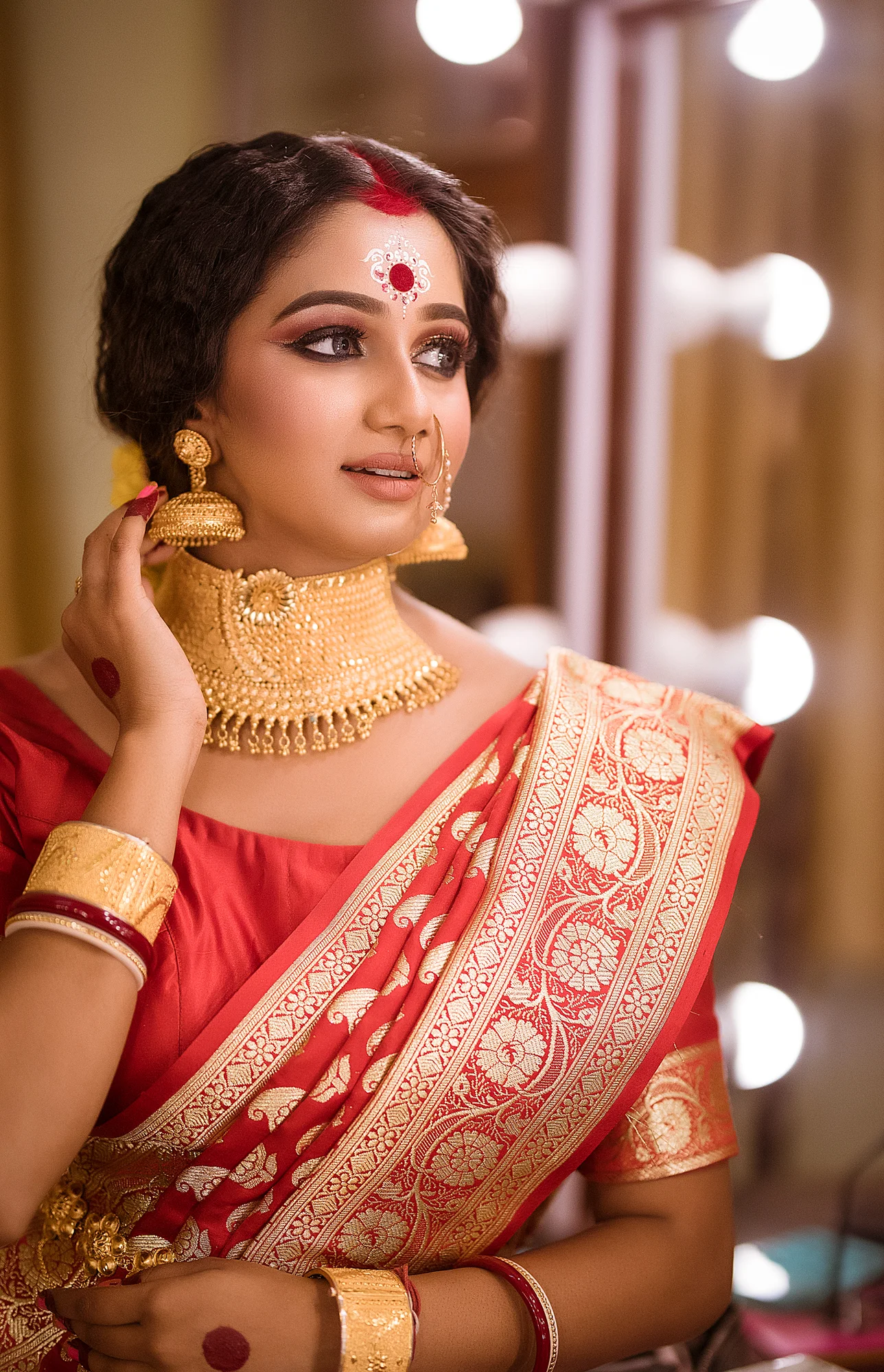A woman dressed in a red and gold traditional Indian saree, adorned with gold jewelry including a choker necklace, earrings, bangles, and a nose ring, standing in front of a mirror with bright lights.