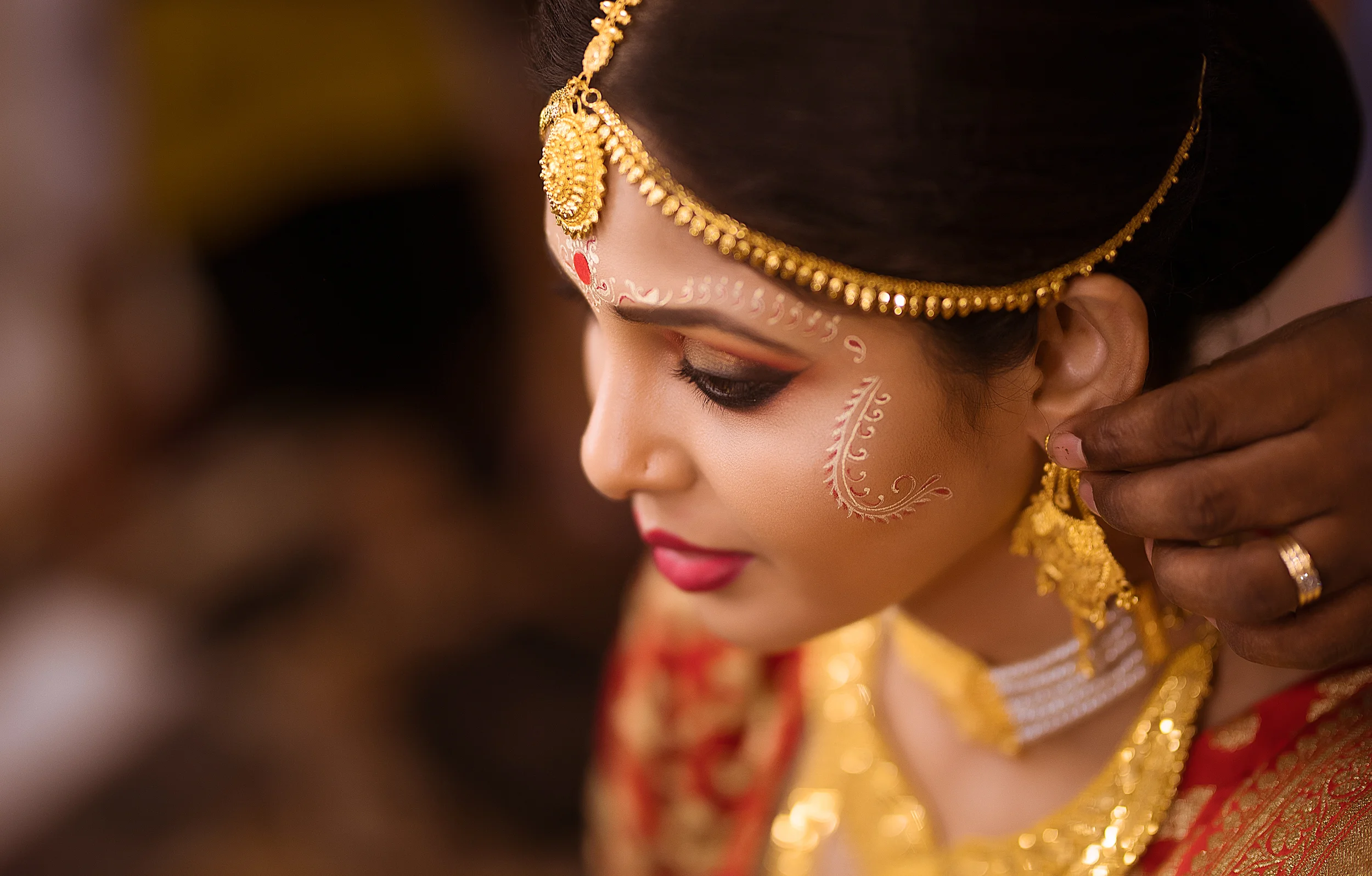 A woman in traditional Indian attire getting her earrings adjusted, with intricate makeup and jewelry, including a headpiece and necklace.