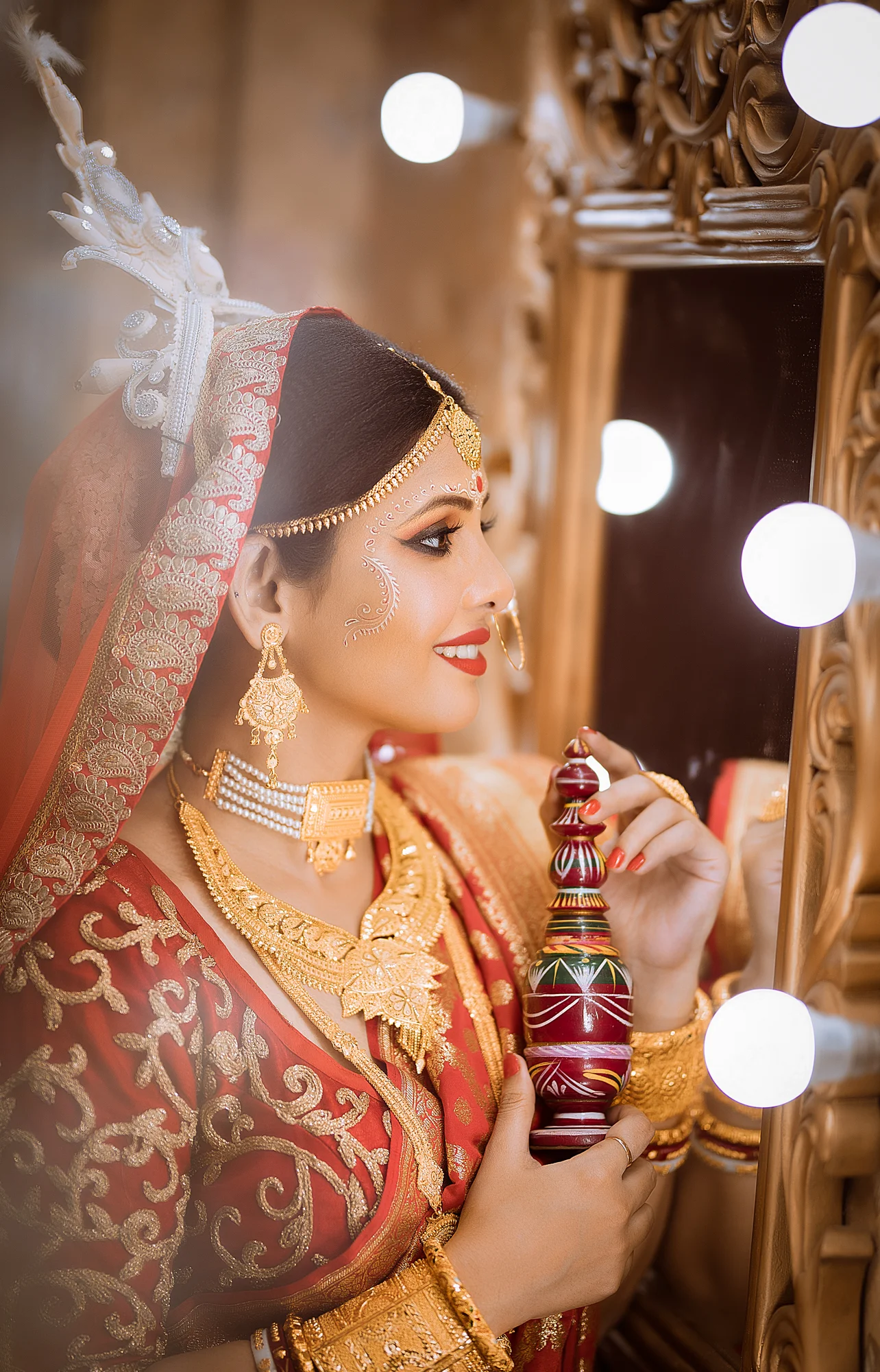 A woman dressed in traditional Indian bridal attire with gold jewelry, a red and gold sari, holding a decorative dandiya stick and looking at her reflection in a mirror surrounded by lights.