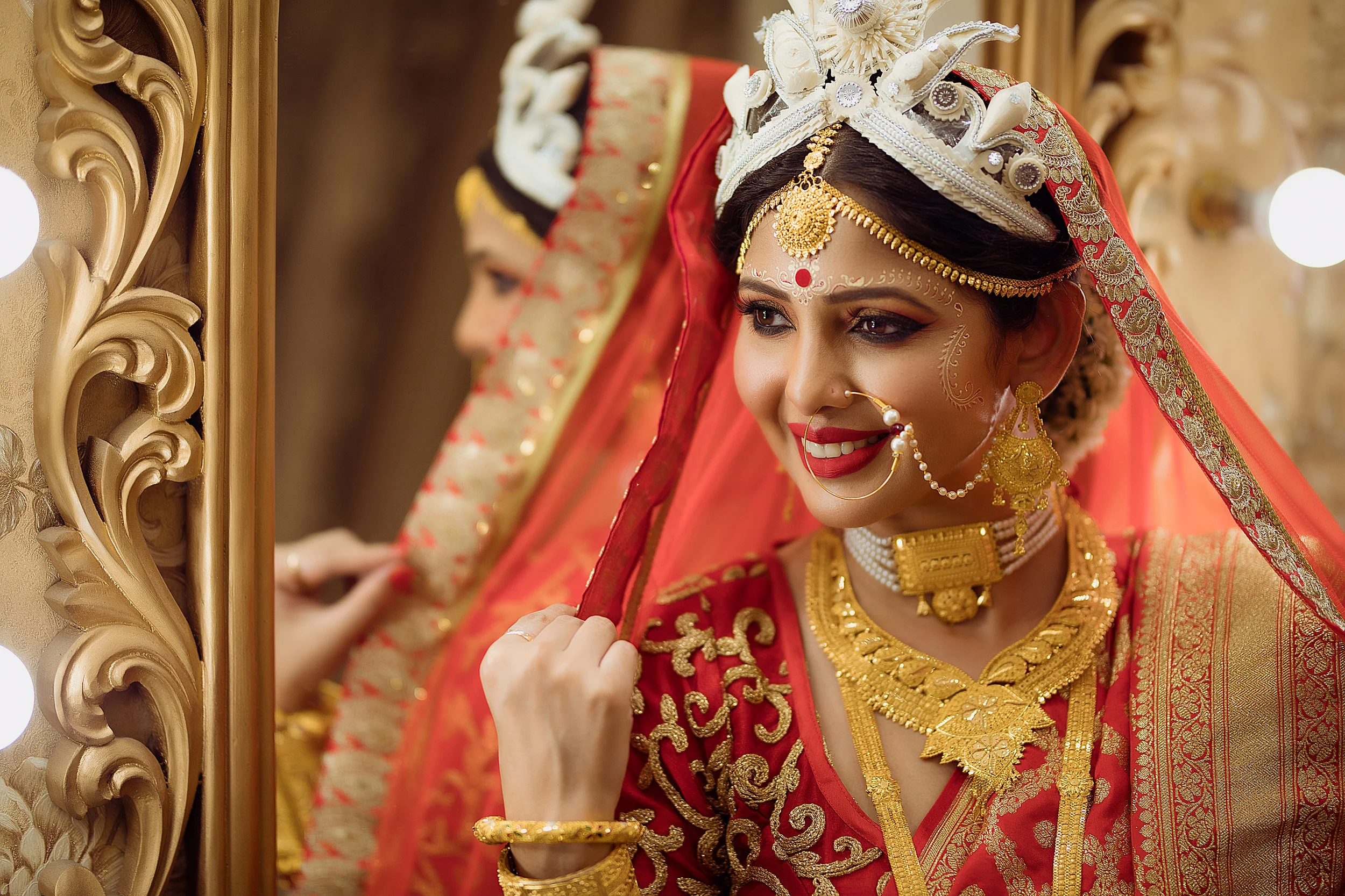 A woman dressed in traditional Indian bridal attire with intricate jewelry, a red and gold saree, and a crown, looking at herself in a mirror with a joyful expression.