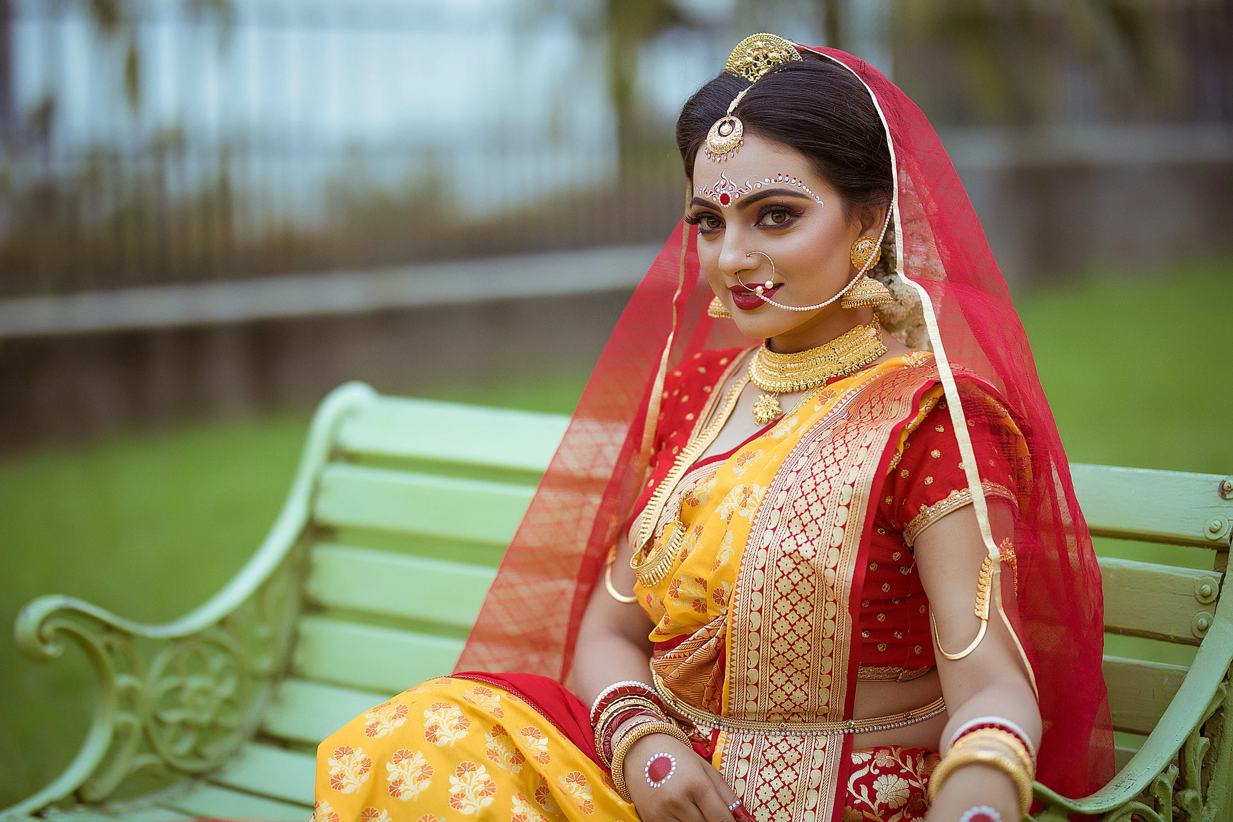 A woman dressed in traditional Indian attire with jewelry, sitting on a green park bench.