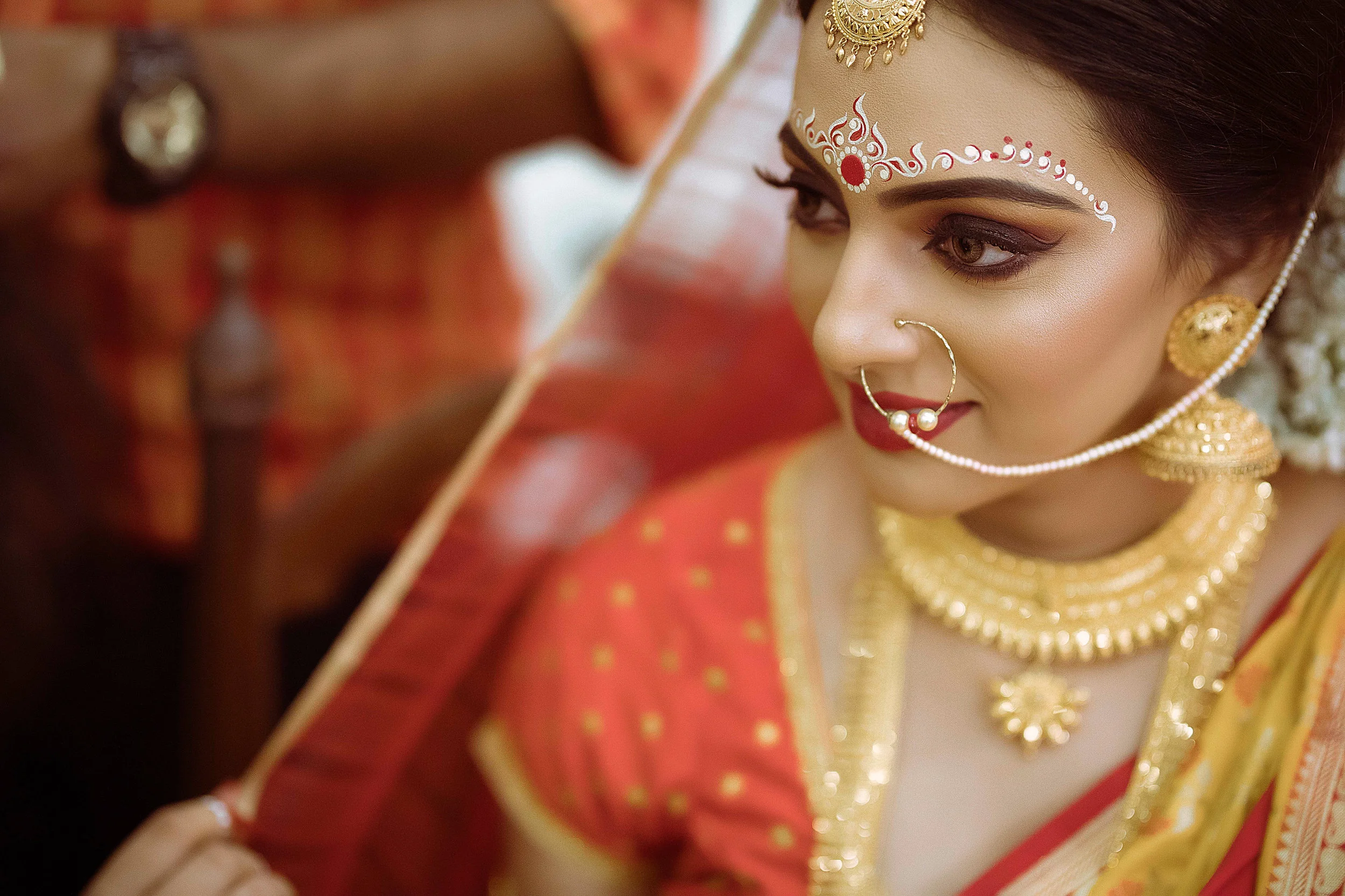 Close-up of an Indian bride adorned with gold jewelry, including a nose ring, earrings, necklace, and headpiece, wearing a red and gold traditional outfit, with intricate forehead and face makeup.