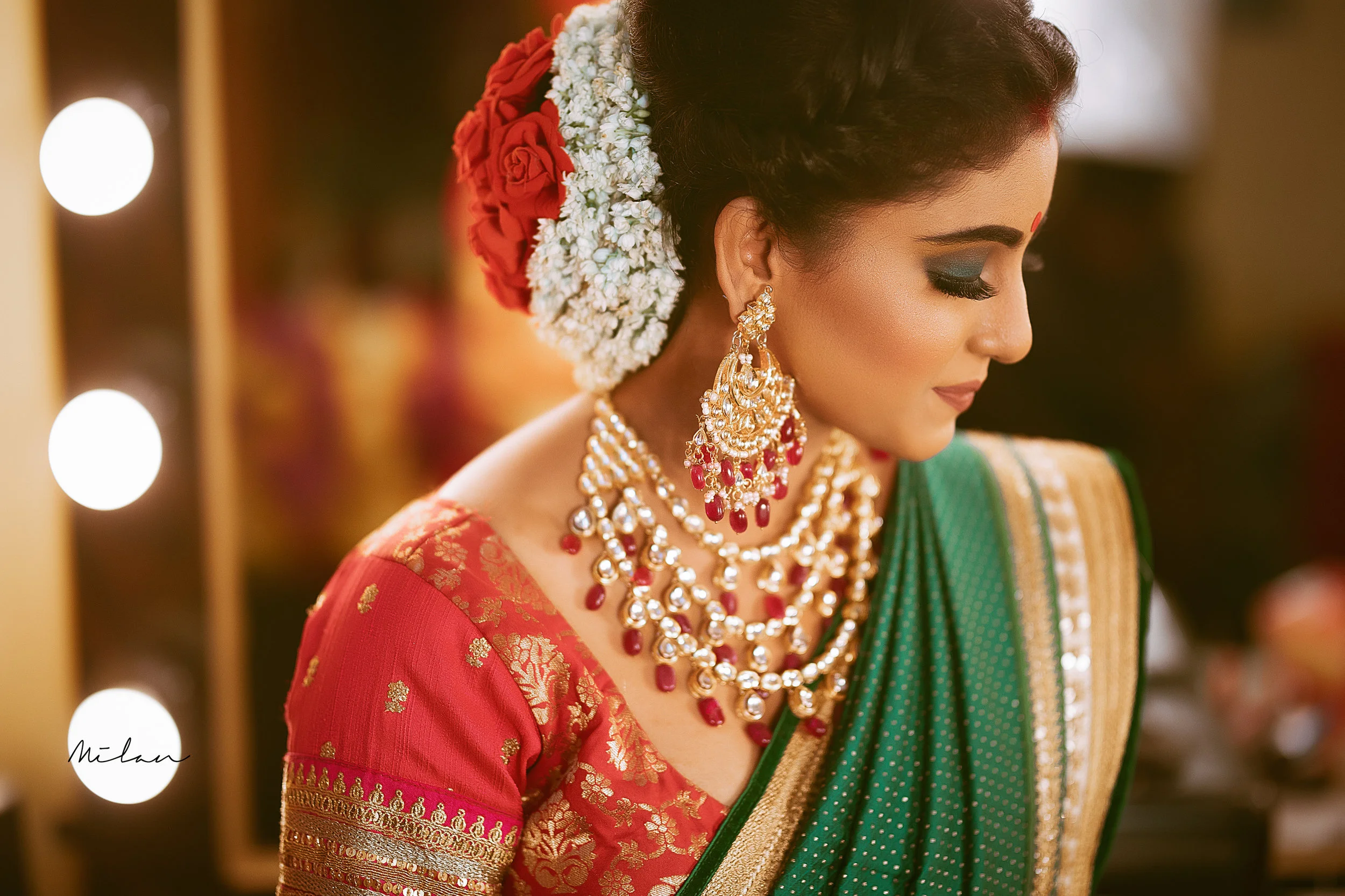 Close-up of an Indian woman dressed in traditional red and green saree with gold embroidery, wearing elaborate gold and red jewelry, including earrings and a layered necklace, with her hair adorned with red and white flowers.