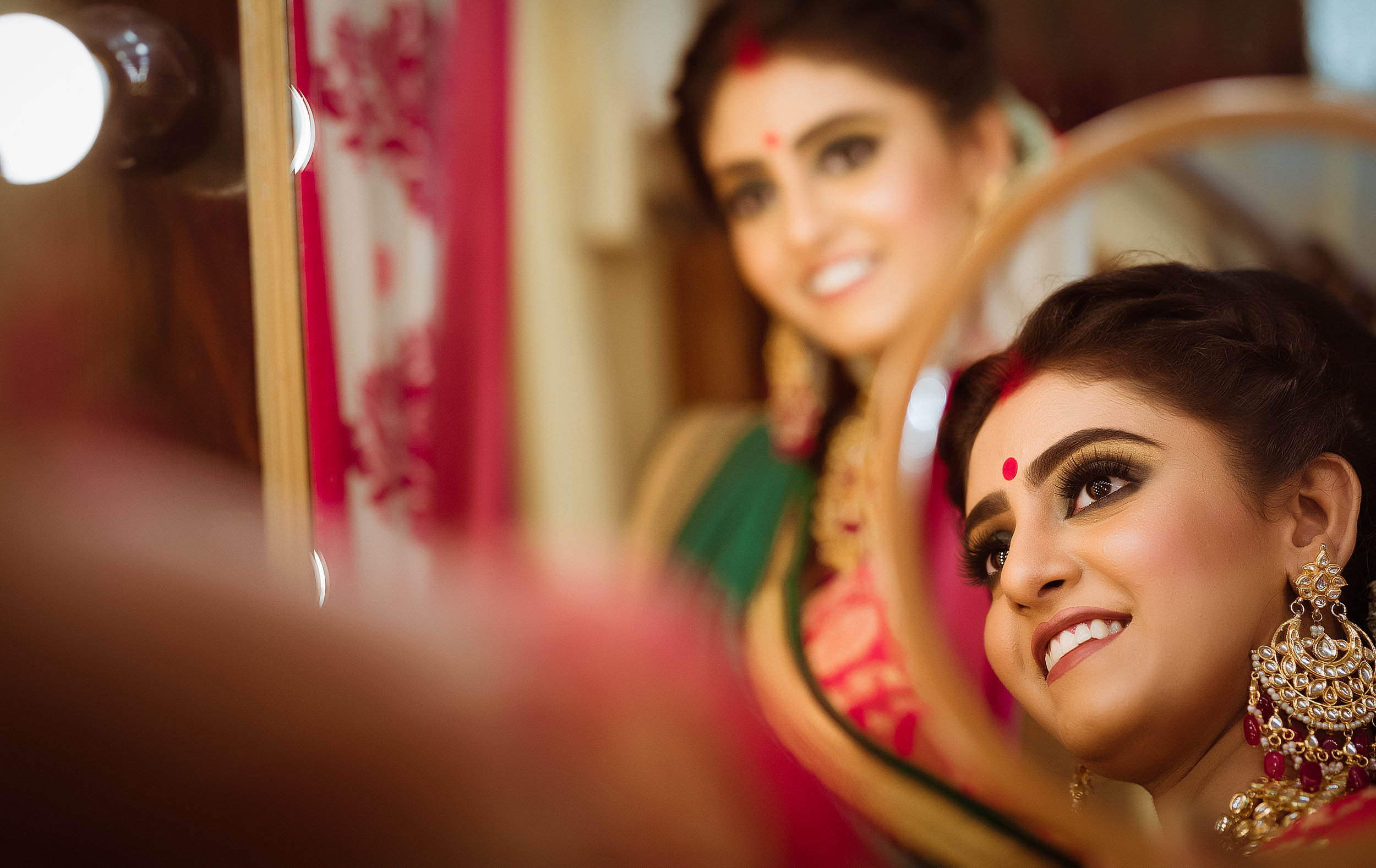 A woman with traditional Indian jewelry and makeup looking into a mirror, smiling.