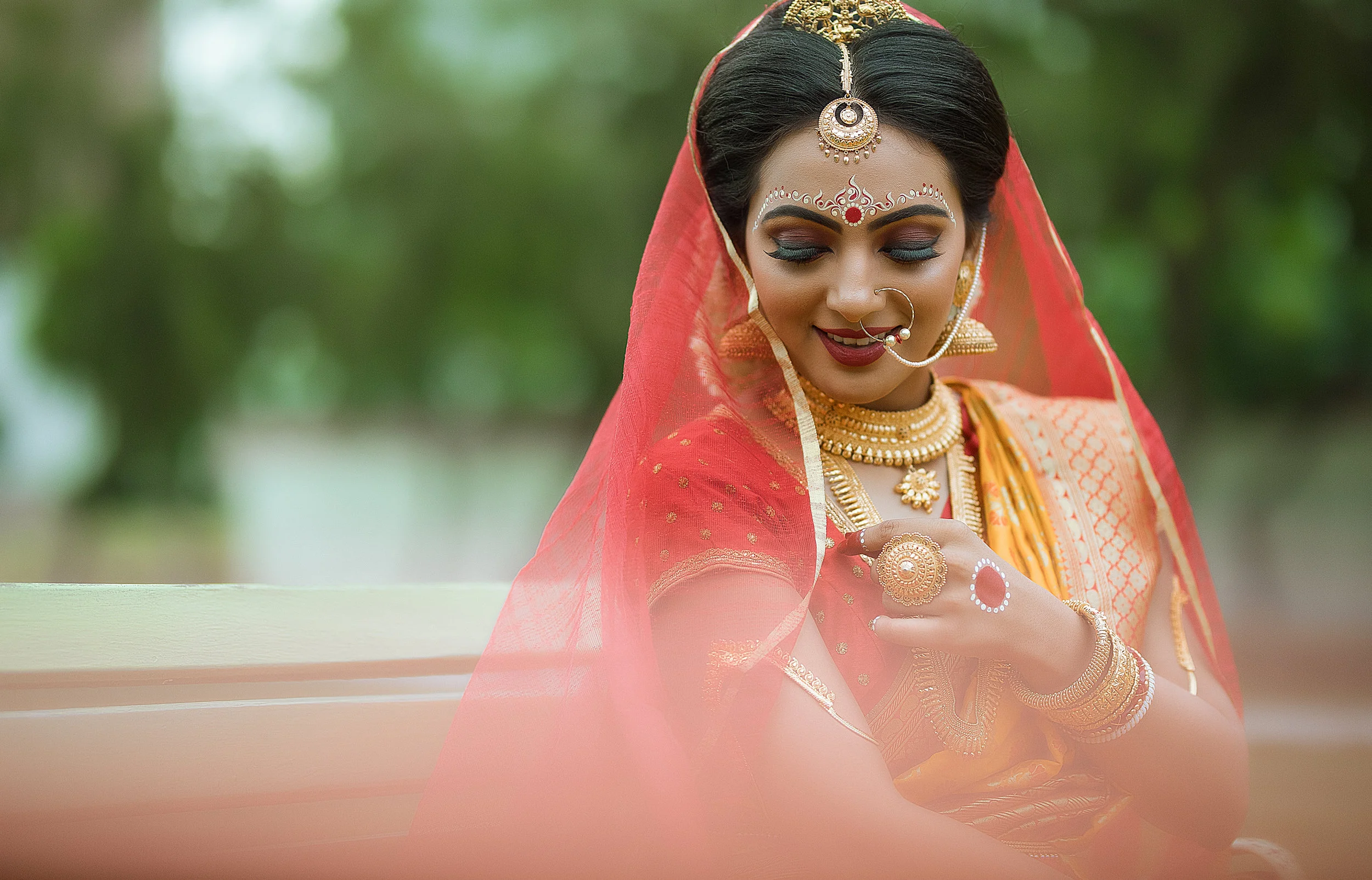 A woman dressed in traditional Indian attire, wearing gold jewelry and a red saree, sits outdoors with a blurred green background. She is smiling with her eyes closed, adorned with decorative makeup and a nose ring.