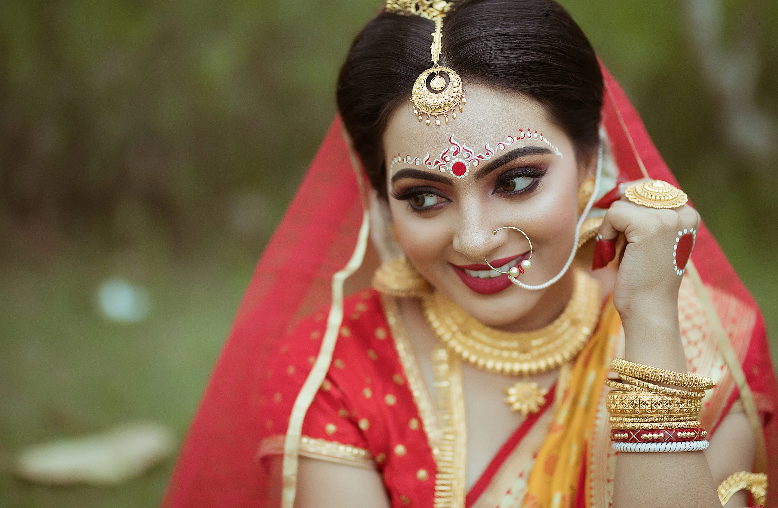 A woman dressed in traditional Indian attire with gold jewelry and makeup, wearing a red and yellow saree, outdoors.