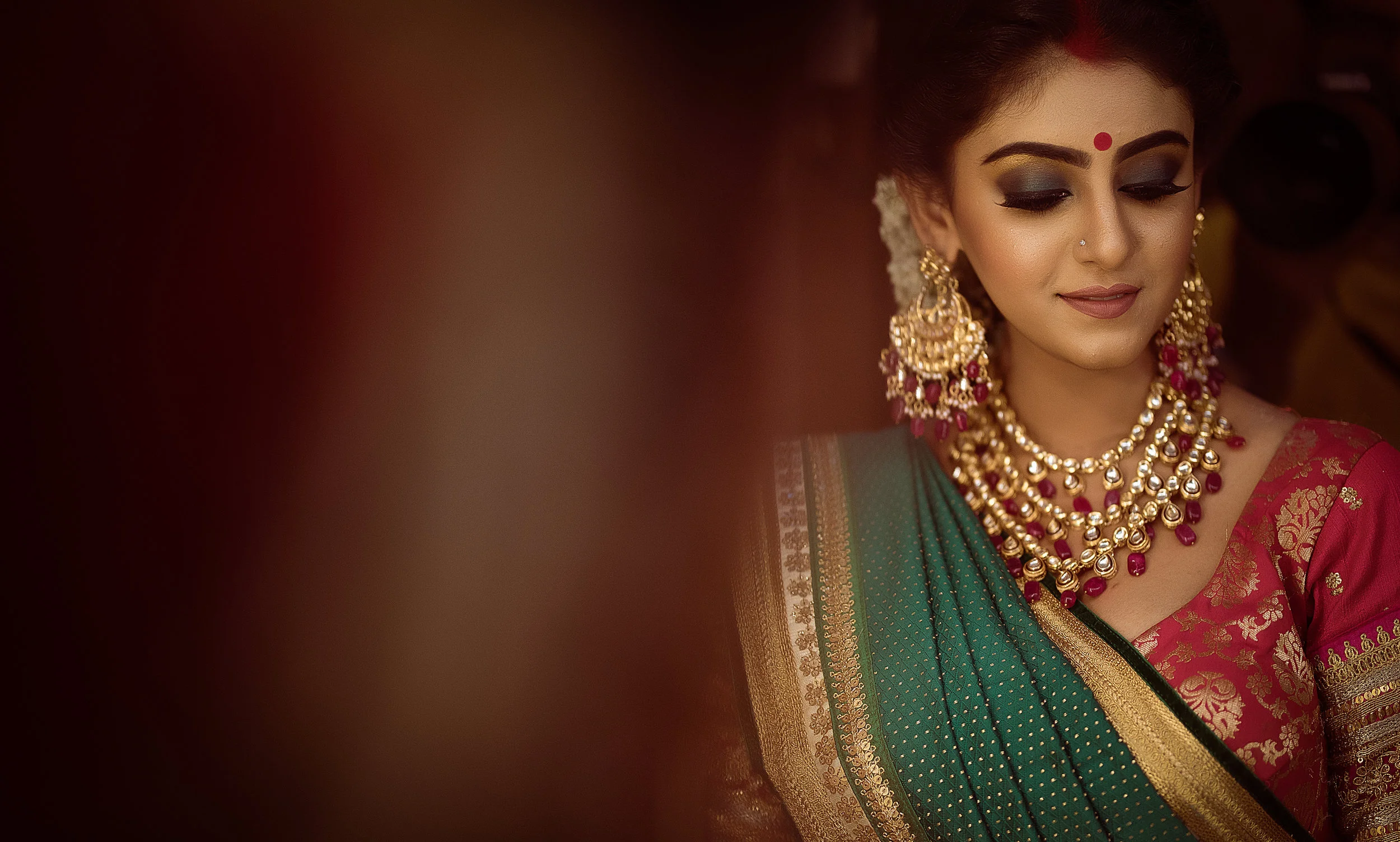 A woman dressed in traditional Indian attire, wearing a teal and gold sari, with elaborate gold and ruby jewelry, and a red bindi on her forehead, her eyes closed and smiling softly.
