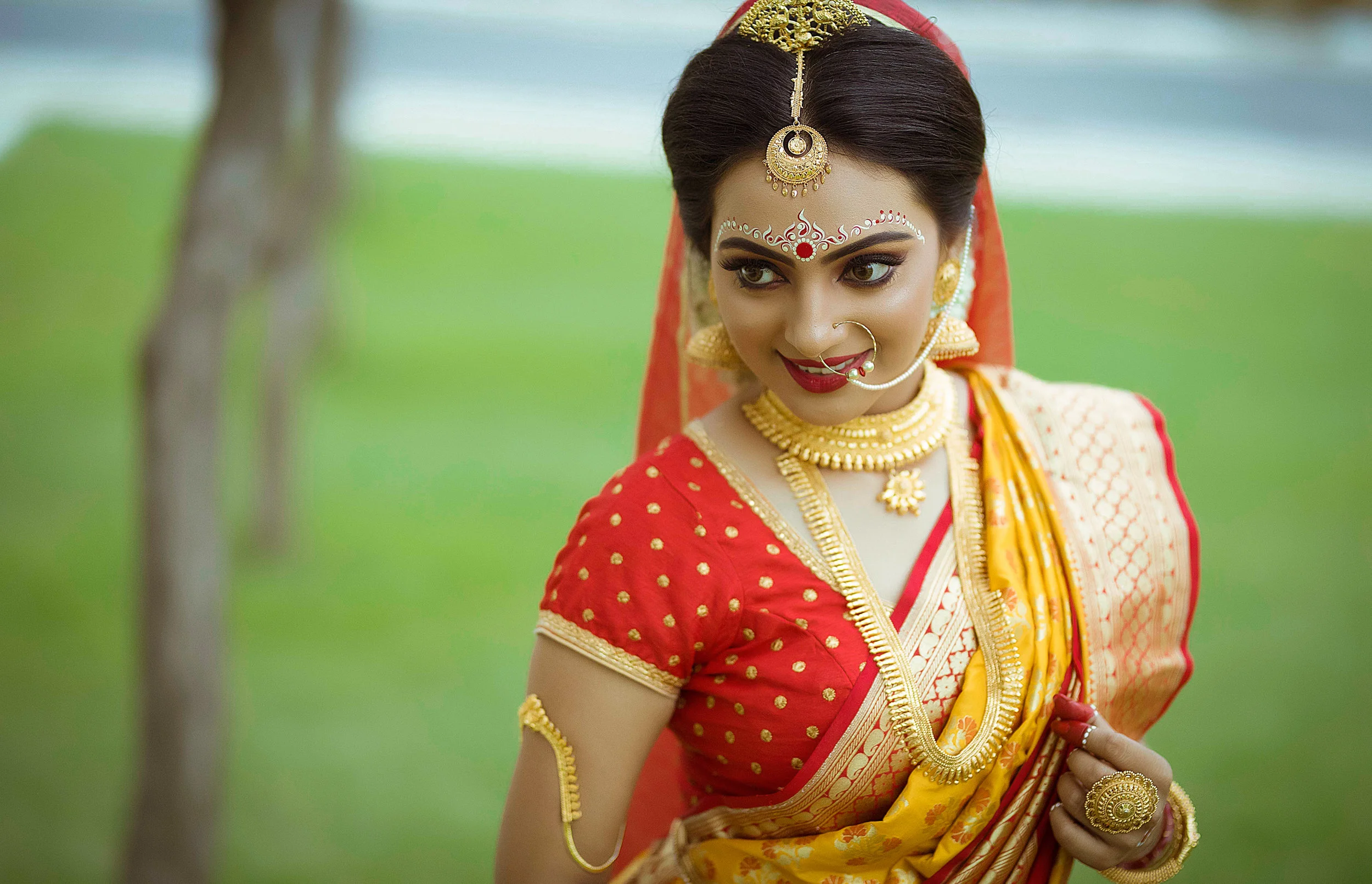 A woman dressed in traditional Indian attire, wearing a red and yellow saree with gold jewelry and decorative accessories, standing outdoors with a blurred green background.