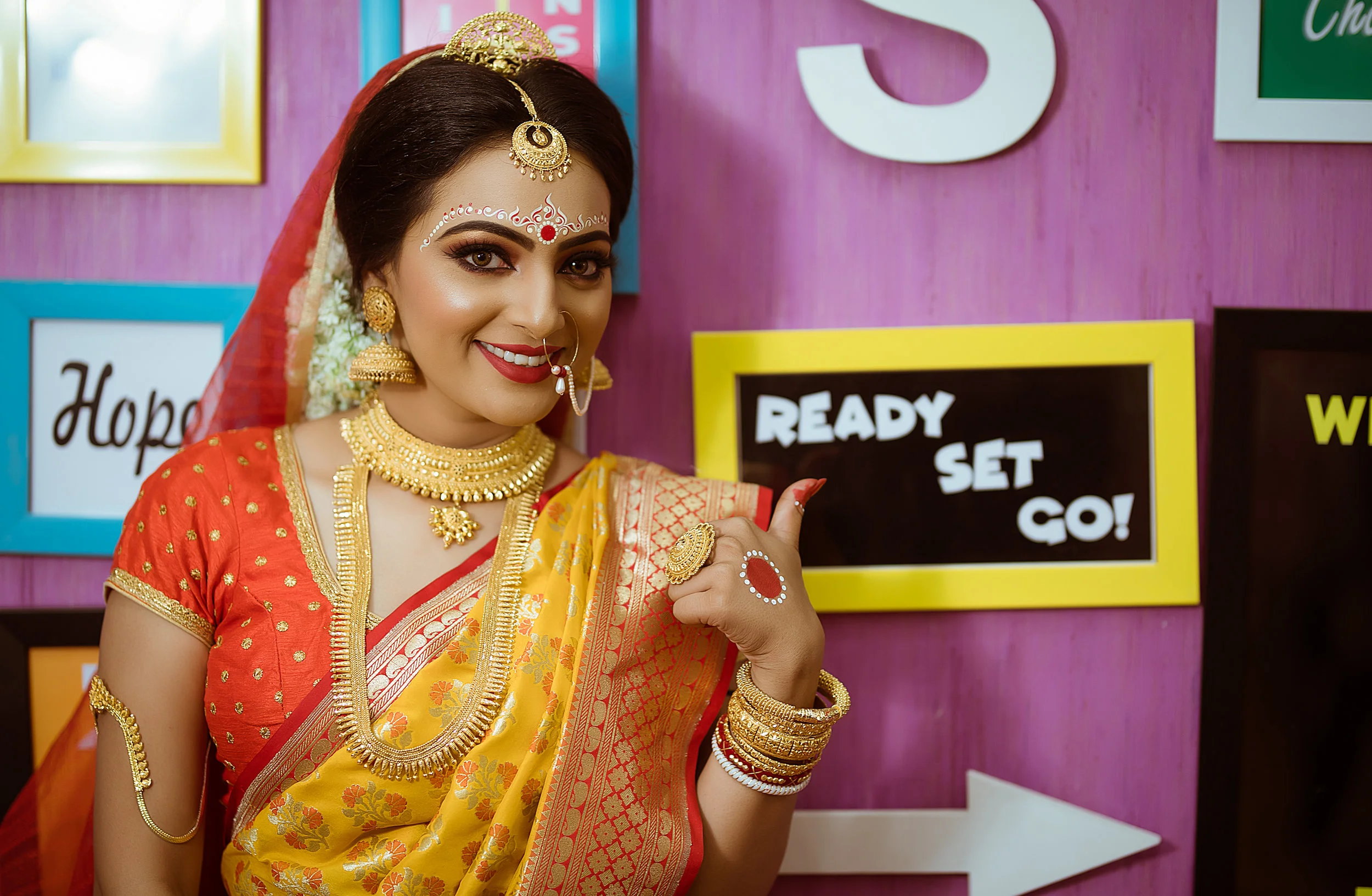 A woman dressed in traditional Indian attire, wearing a red and yellow saree and gold jewelry, standing in front of colorful signs, smiling and pointing to a sign that says "Ready Set Go!"