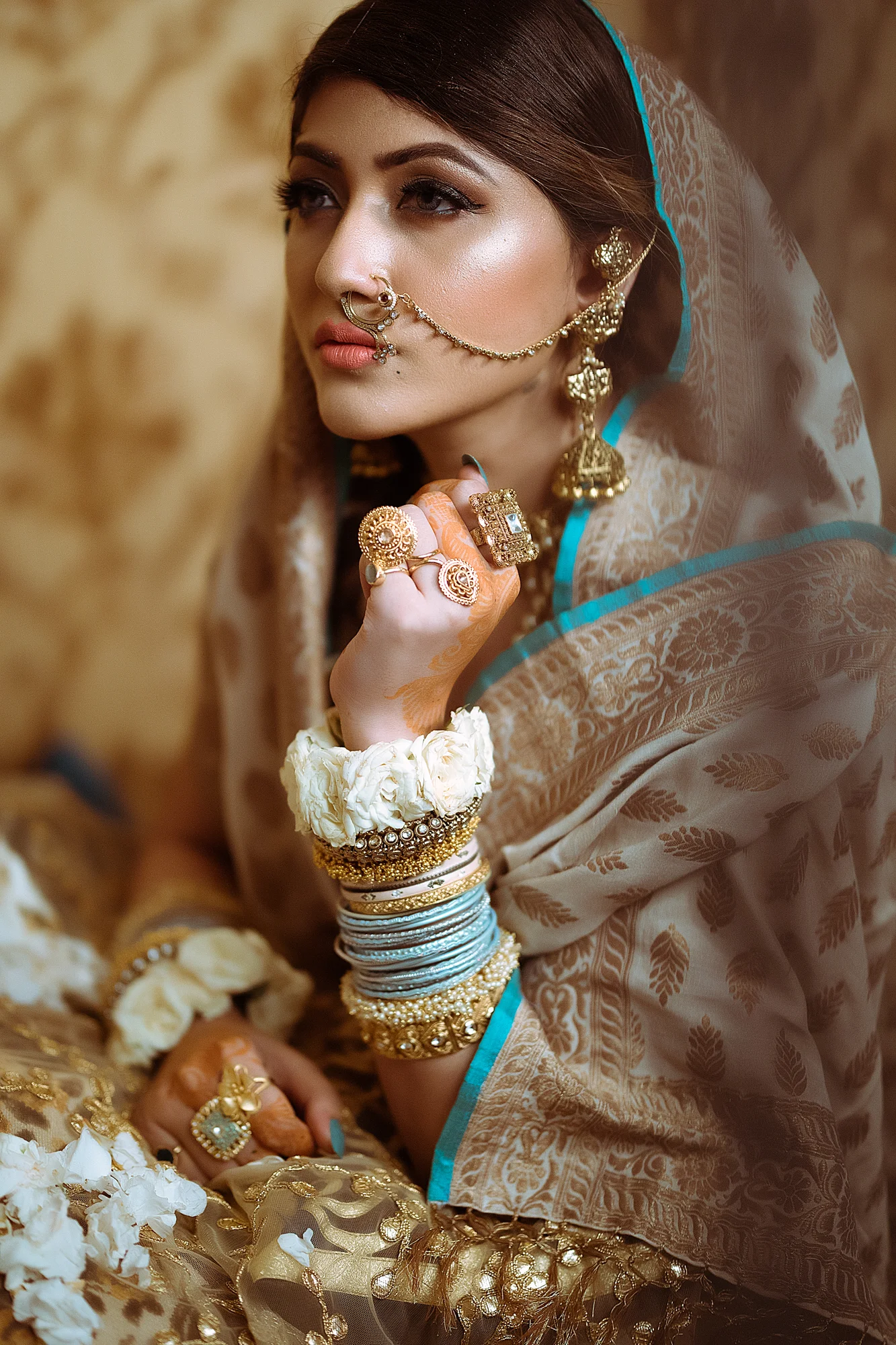 A woman dressed in traditional Indian attire, adorned with gold jewelry, several bangles, and rings, sitting with flowers around her.