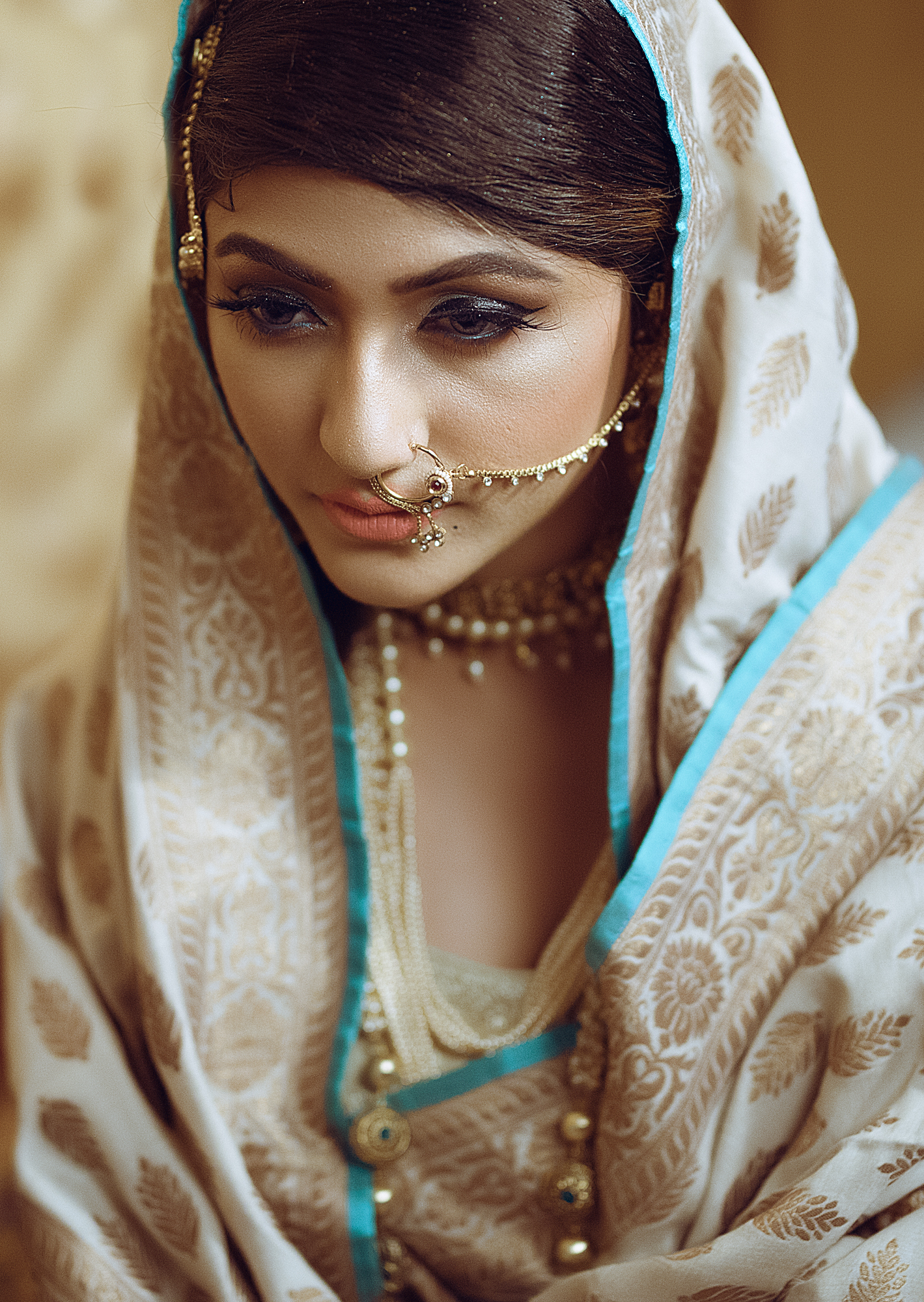 A woman dressed in traditional Indian attire, wearing a beige and gold saree with a blue border, adorned with gold jewelry, including a nose ring, earrings, and necklaces, with a serious and contemplative expression.