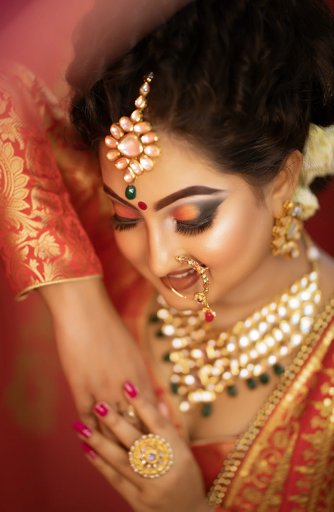 Close-up of a woman dressed in traditional Indian attire with elaborate gold jewelry, including earrings, necklace, and nose ring, with her eyes closed, wearing vibrant makeup, and adorned with a red bindi and decorative embellishments on her forehea