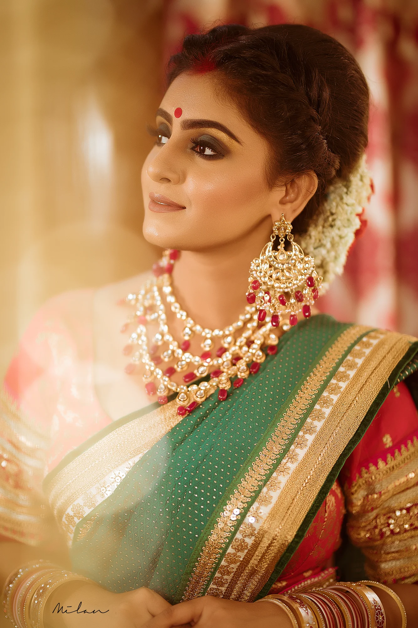 A woman dressed in traditional Indian attire, wearing a green saree with gold accents, adorned with elaborate gold and red jewelry, including earrings, necklaces, and bangles, has her hair decorated with white flowers and is smiling softly, looking t