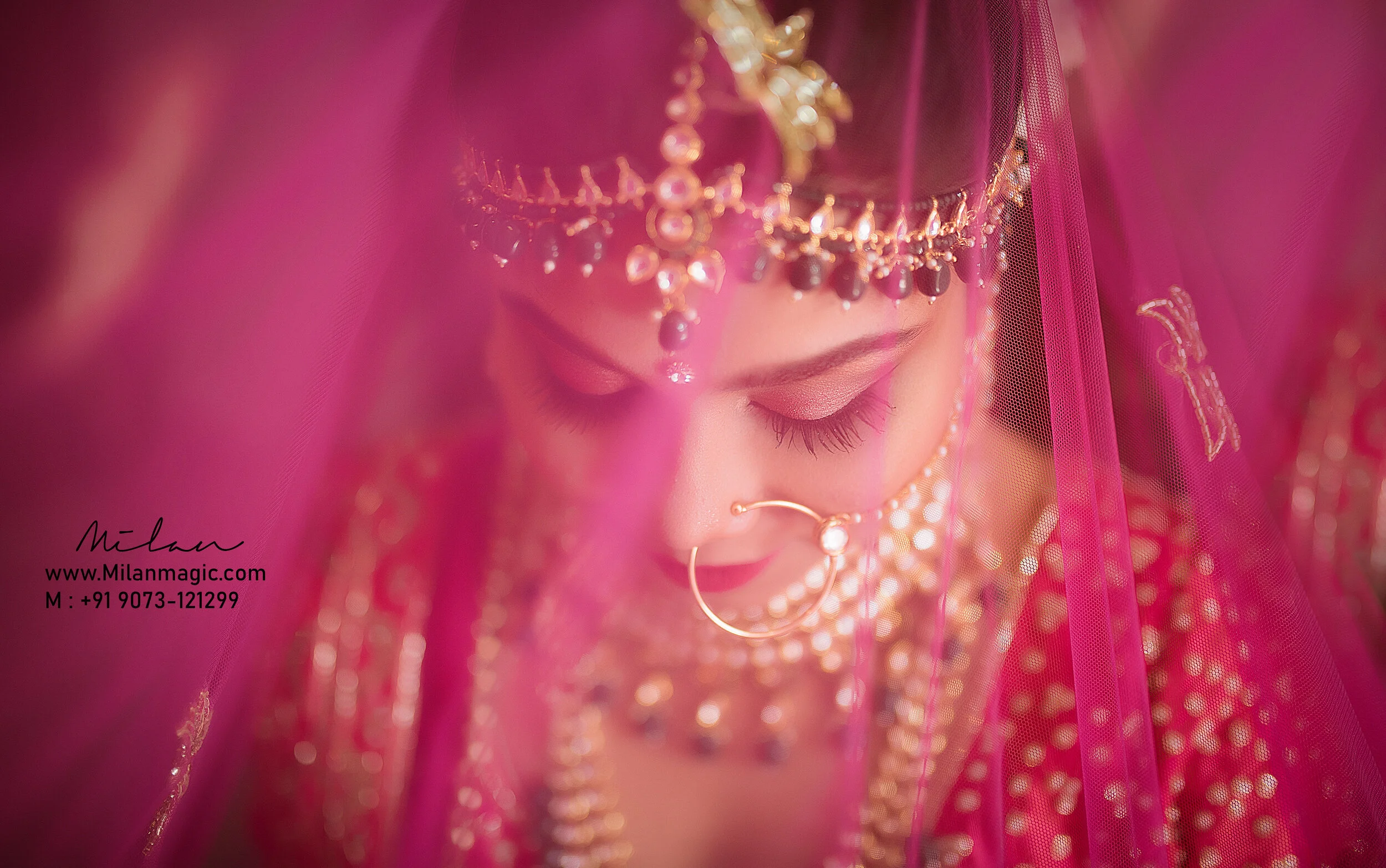 Close-up of a bride in traditional Indian bridal attire, wearing a gold and gem jewelry including a nose ring, necklace, and headpiece with a pink veil covering her face and head.