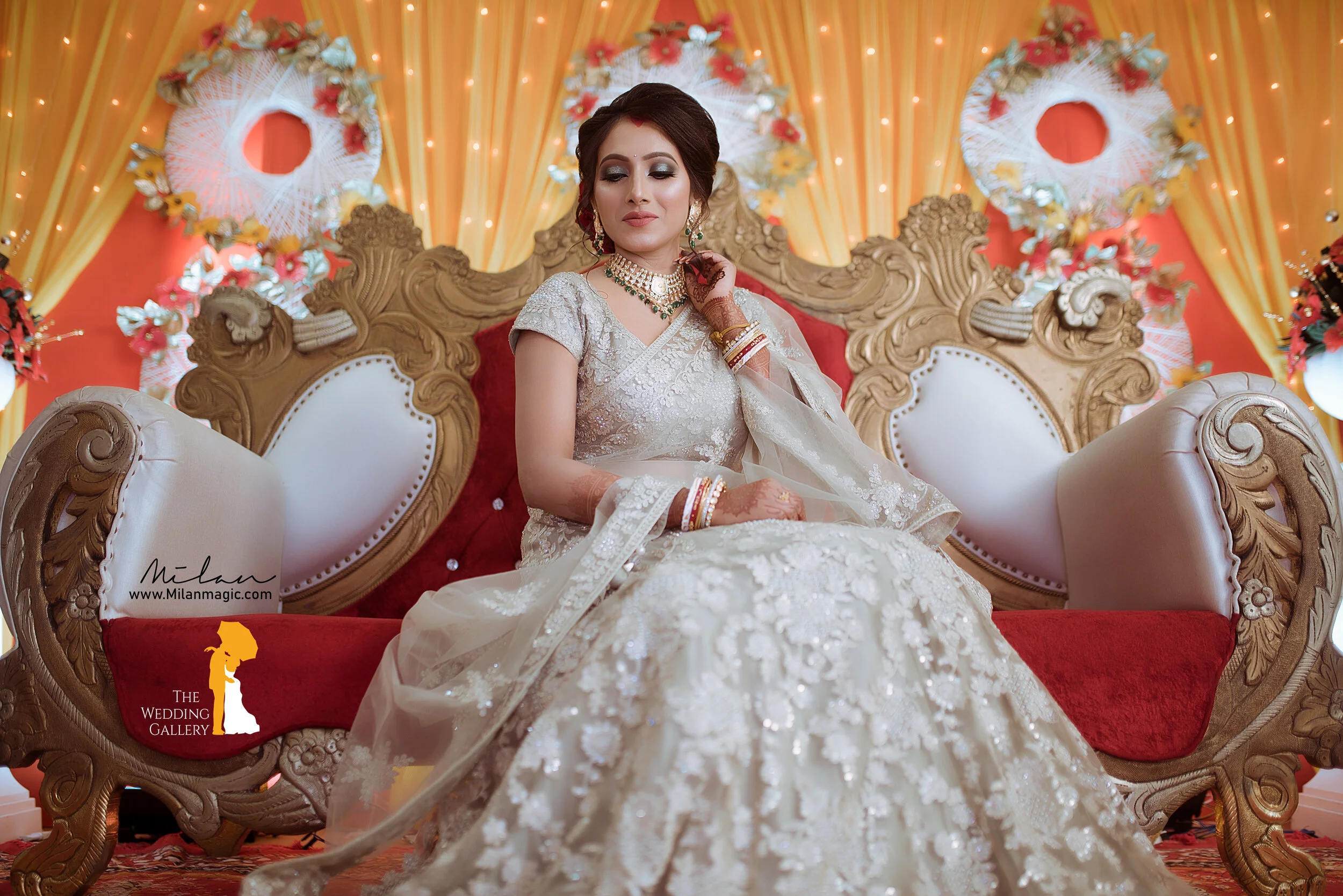 A bride sitting on an ornate, carved wooden sofa with white and red upholstery, wearing a cream-colored embroidered wedding dress and jewelry, in front of a decorated backdrop with orange and yellow drapes and floral wreaths.