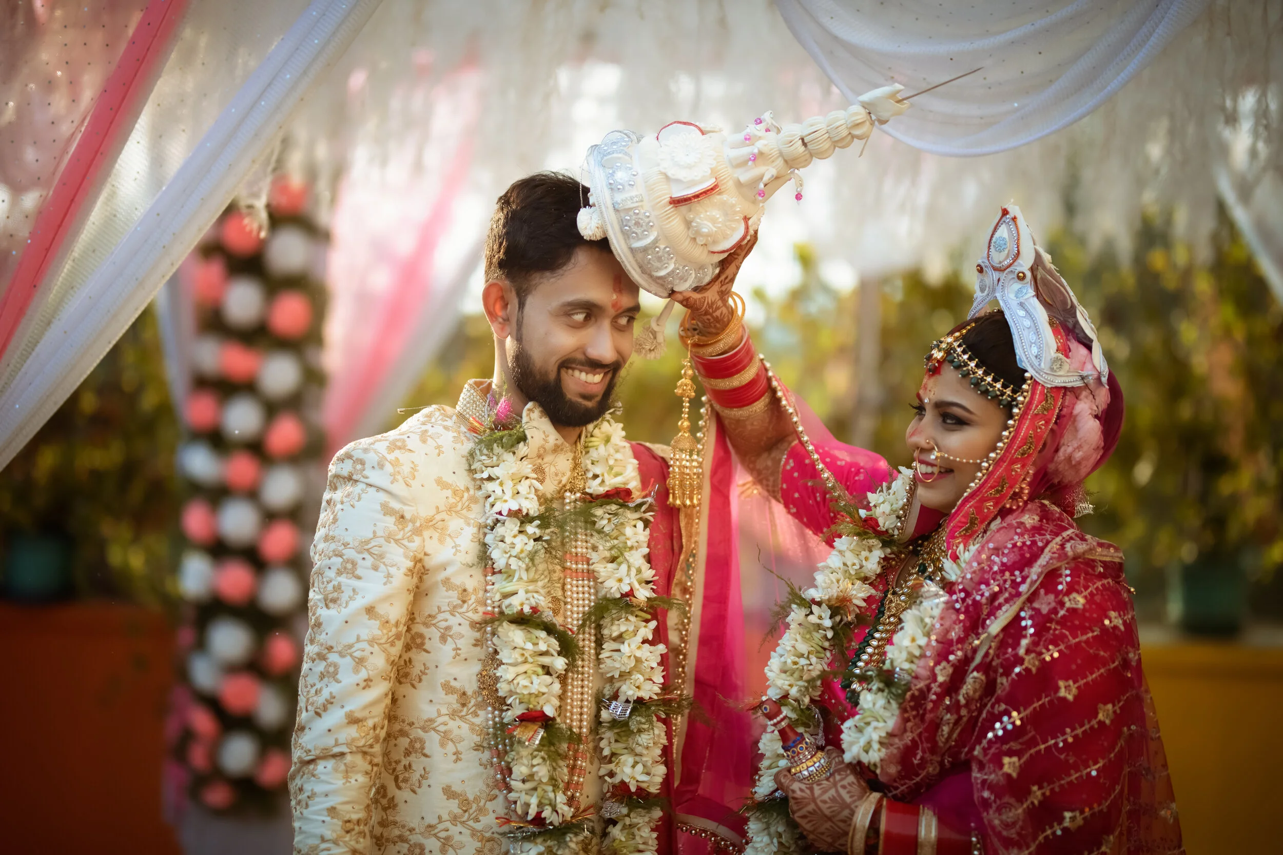 Indian couple during wedding ceremony, the bride is smiling and wearing traditional red saree and jewelry, the groom is smiling and wearing a cream-colored sherwani, an elder woman is pouring water on the groom's head with a decorated pot, in an outd