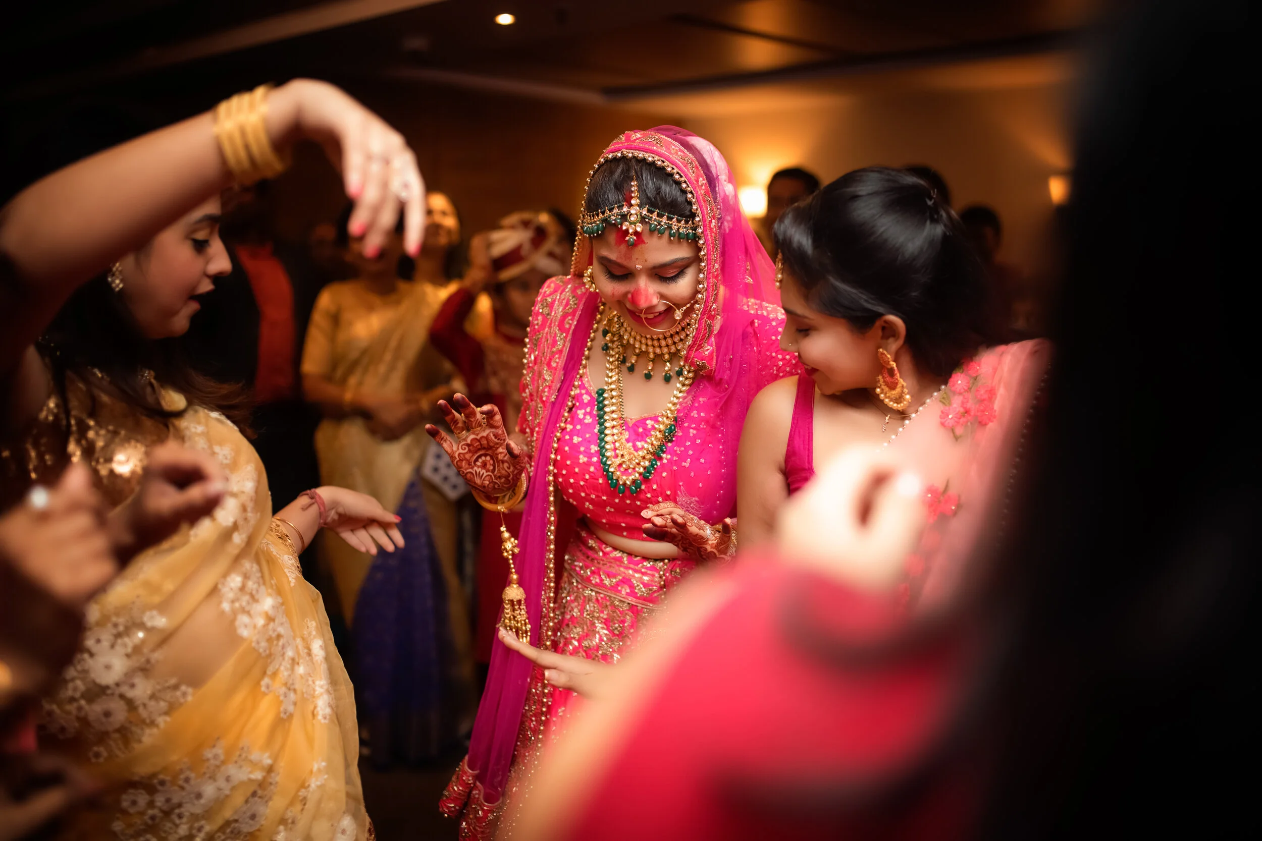 Women celebrating at an Indian wedding, dressed in colorful traditional attire, with the bride wearing pink and gold jewelry.