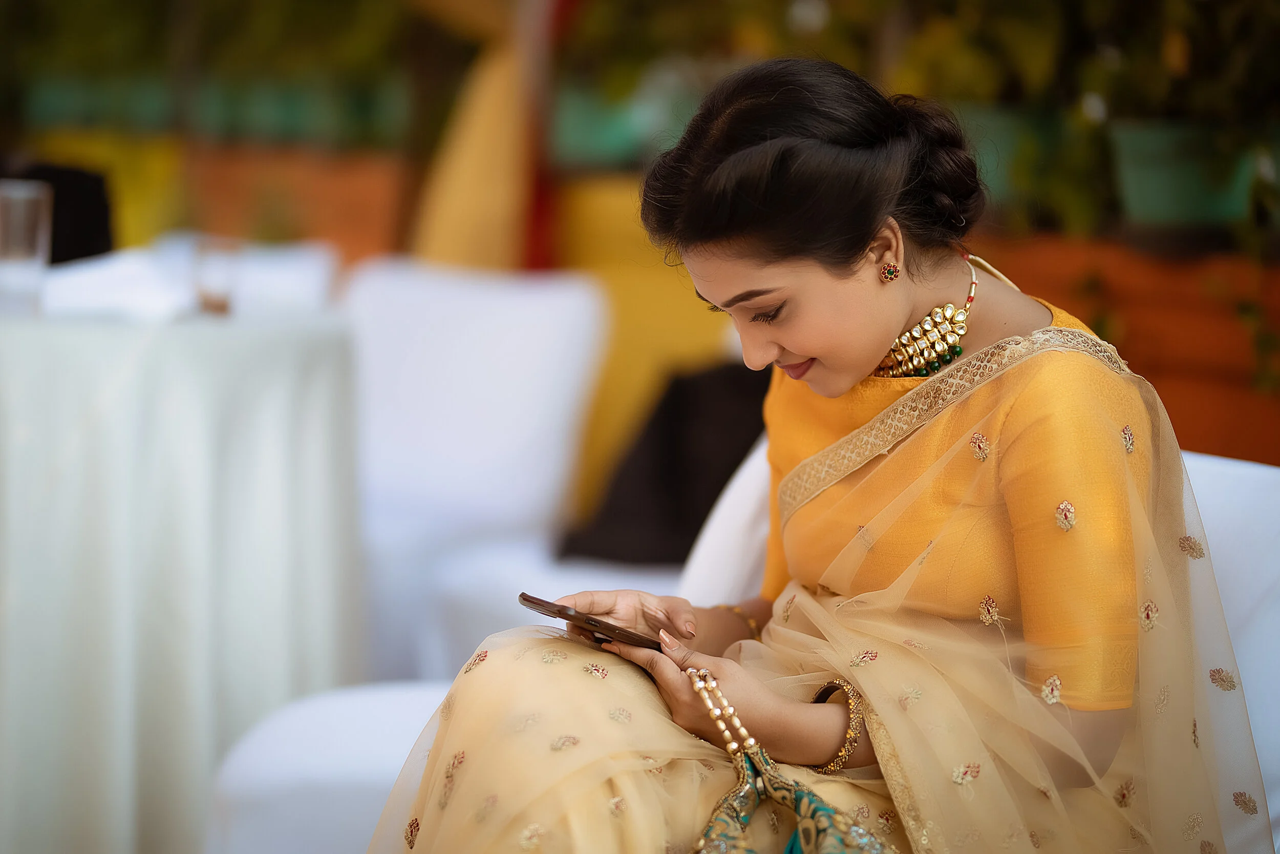 A woman dressed in traditional Indian attire, adorned with jewelry, sitting at an outdoor event, looking at her phone.